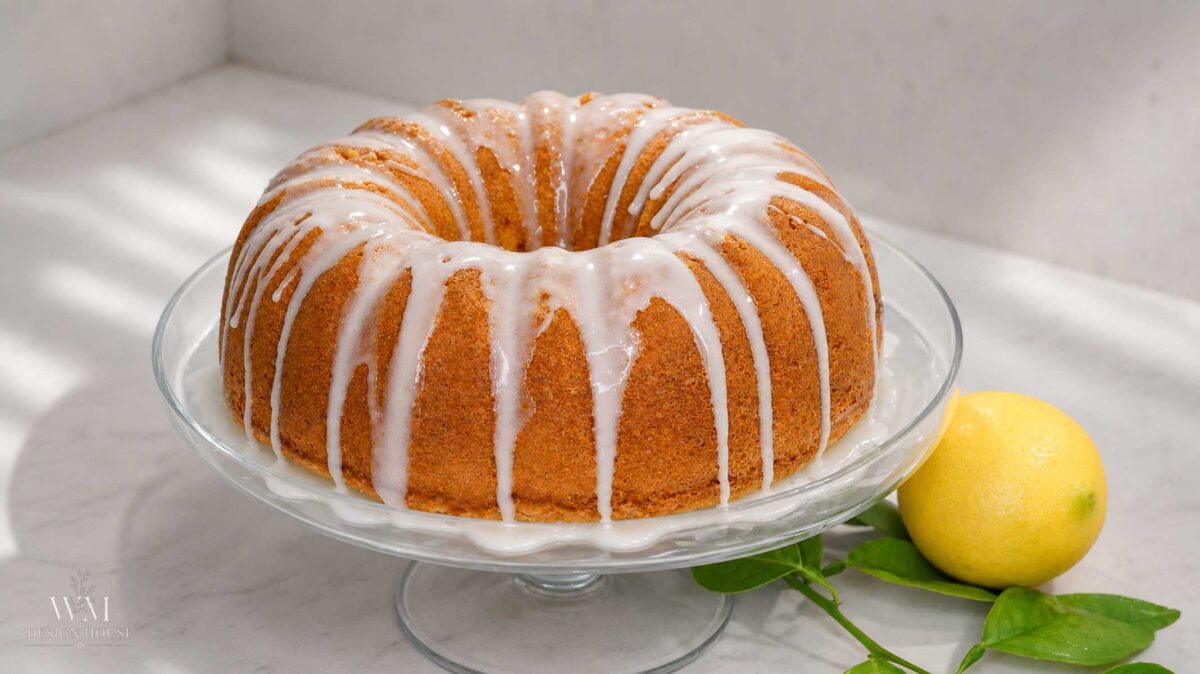 A glazed lemon bundt cake on a footed glass cake stand with a fresh lemon alongside