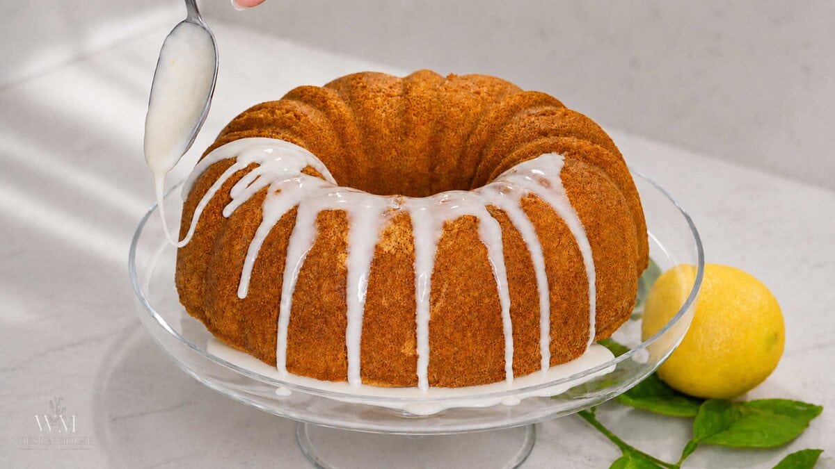 A spoon drizzles white icing over a golden-brown lemon curd bundt cake on a glass stand. A whole lemon and green leaves are placed beside the cake on a white countertop.