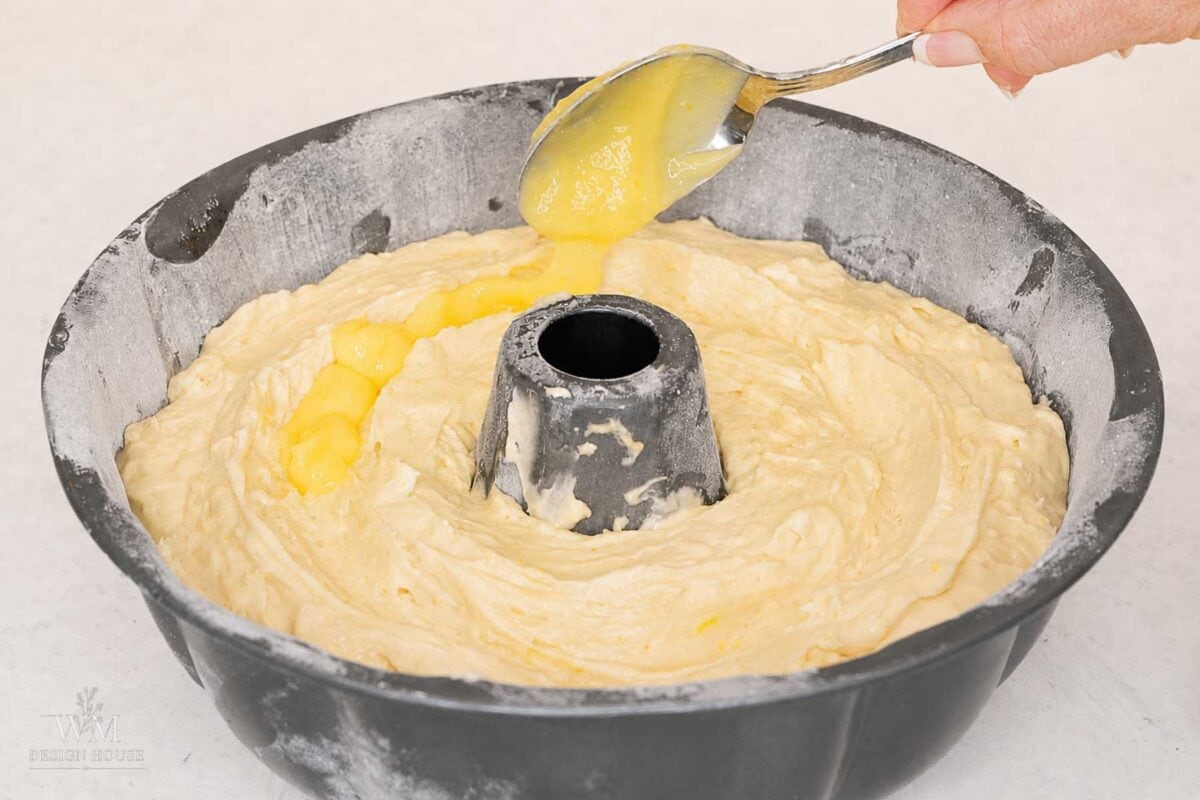 A hand uses a spoon to add yellow lemon filling onto cake batter in a floured bundt pan, preparing to bake a lemon-filled cake.
