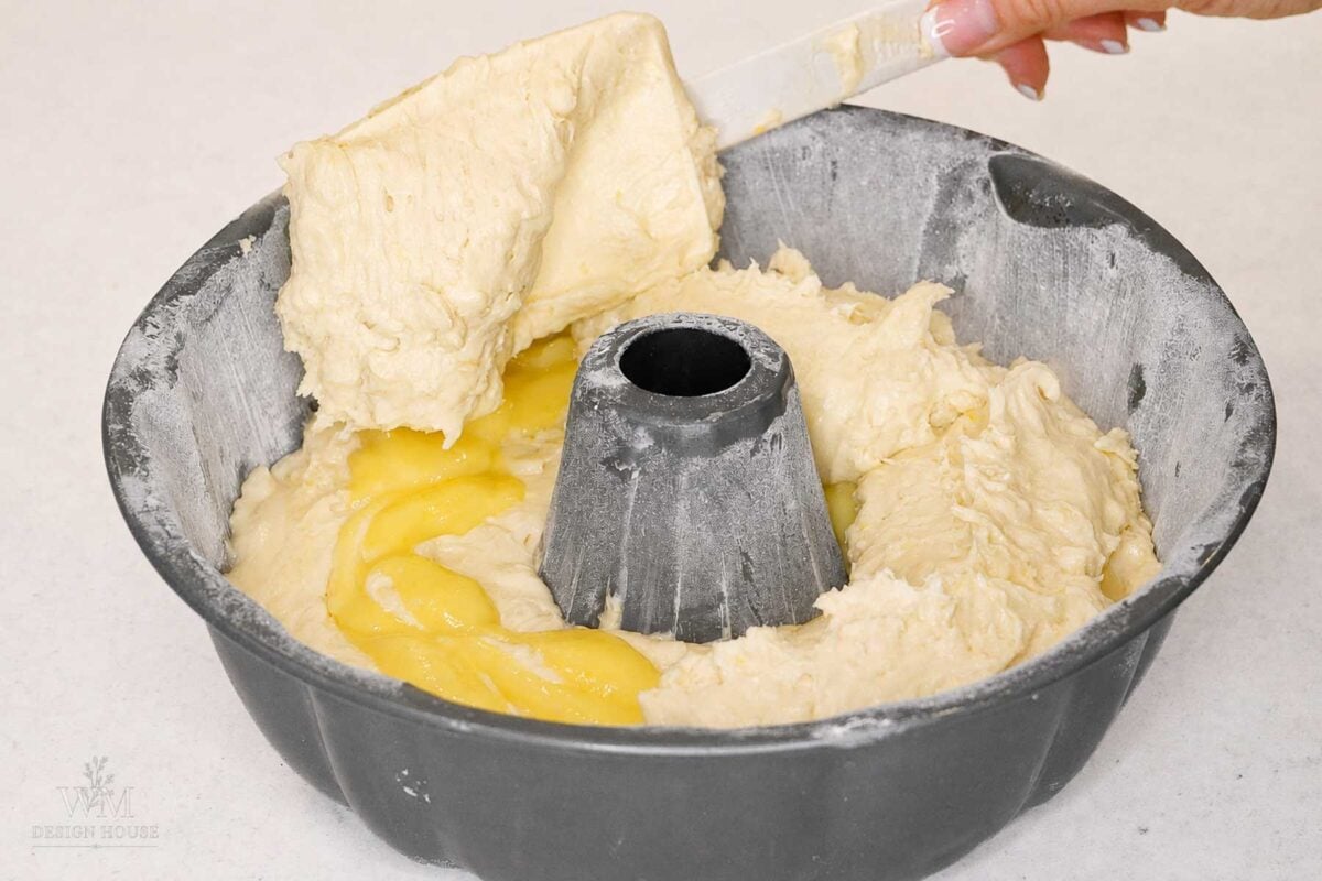 A hand spreads lemon cake batter over a second layer of lemon curd into a floured bundt cake pan, preparing it for baking on a light countertop.