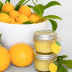 A white bowl of fresh lemons with green leaves sits behind two small jars of lemon curd, each tied with twine and a lemon-shaped tag. Several loose lemons and leafy stems are arranged around the jars.