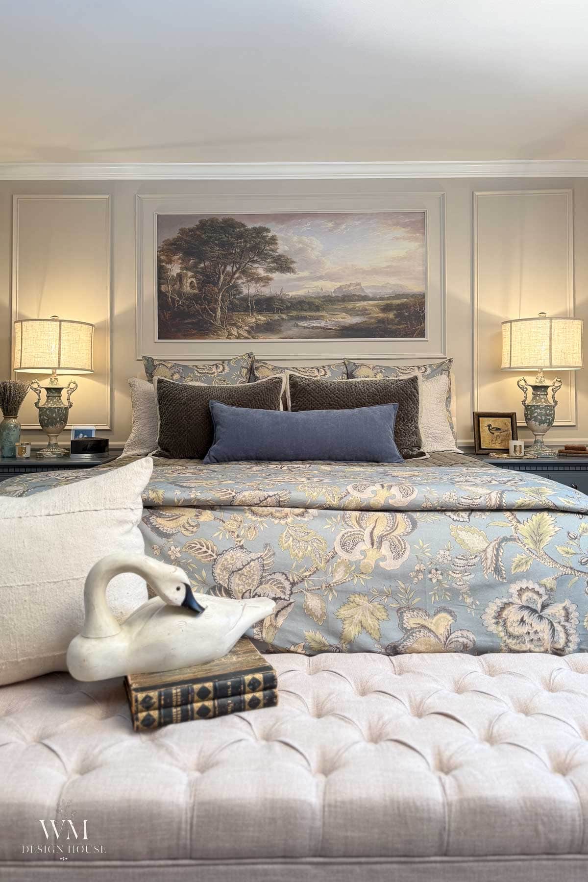 A cozy bedroom with a patterned bedspread, decorative pillows, two lamps on nightstands, a landscape painting above the bed, and a tufted bench with a ceramic swan and vintage books.