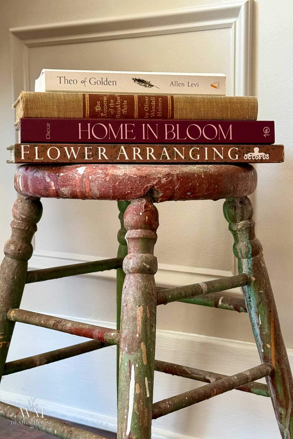 Four books, including “Home in Bloom” and “Flower Arranging,” stacked on a weathered, multicolored wooden stool against a white wall.
