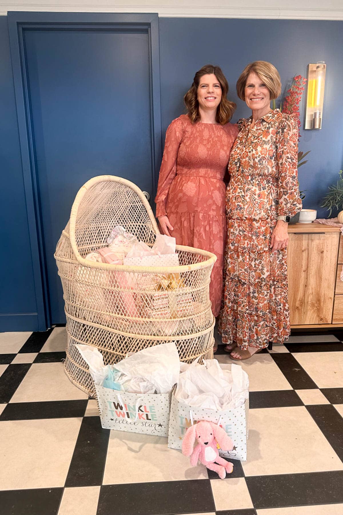 Two women in long dresses stand smiling beside a wicker bassinet filled with gifts. More gift bags, including one with a pink stuffed bunny, are on the black-and-white tiled floor in front of them.