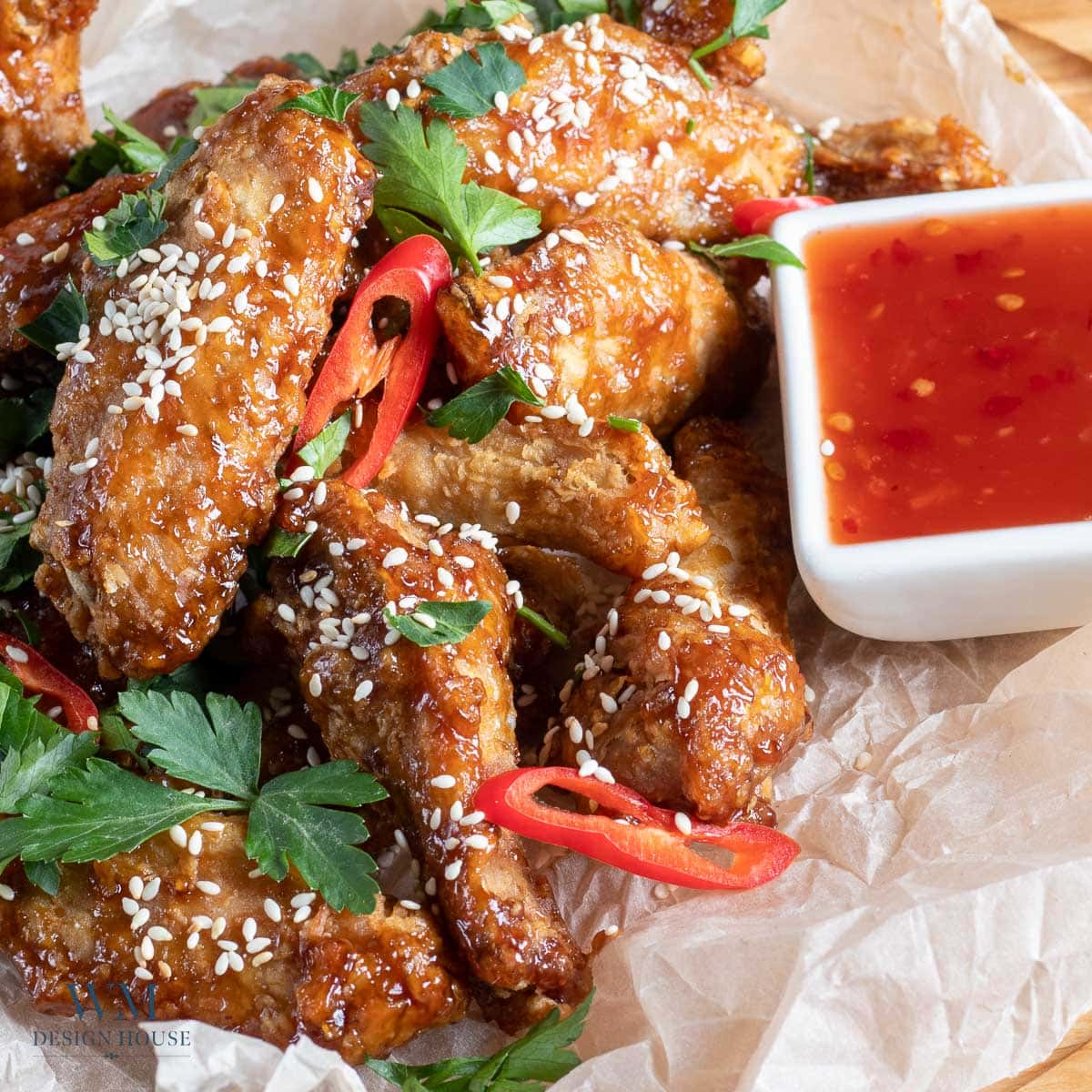 A pile of glazed chicken wings garnished with sesame seeds, parsley, and sliced red chili, served with a square dish of red dipping sauce on crumpled parchment paper.