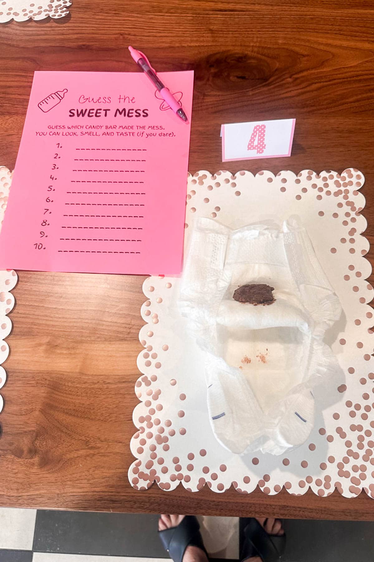 A diaper with brown candy is on a decorated placemat next to a pink “Guess the Sweet Mess” game sheet, a pen, and a numbered card on a wooden table.