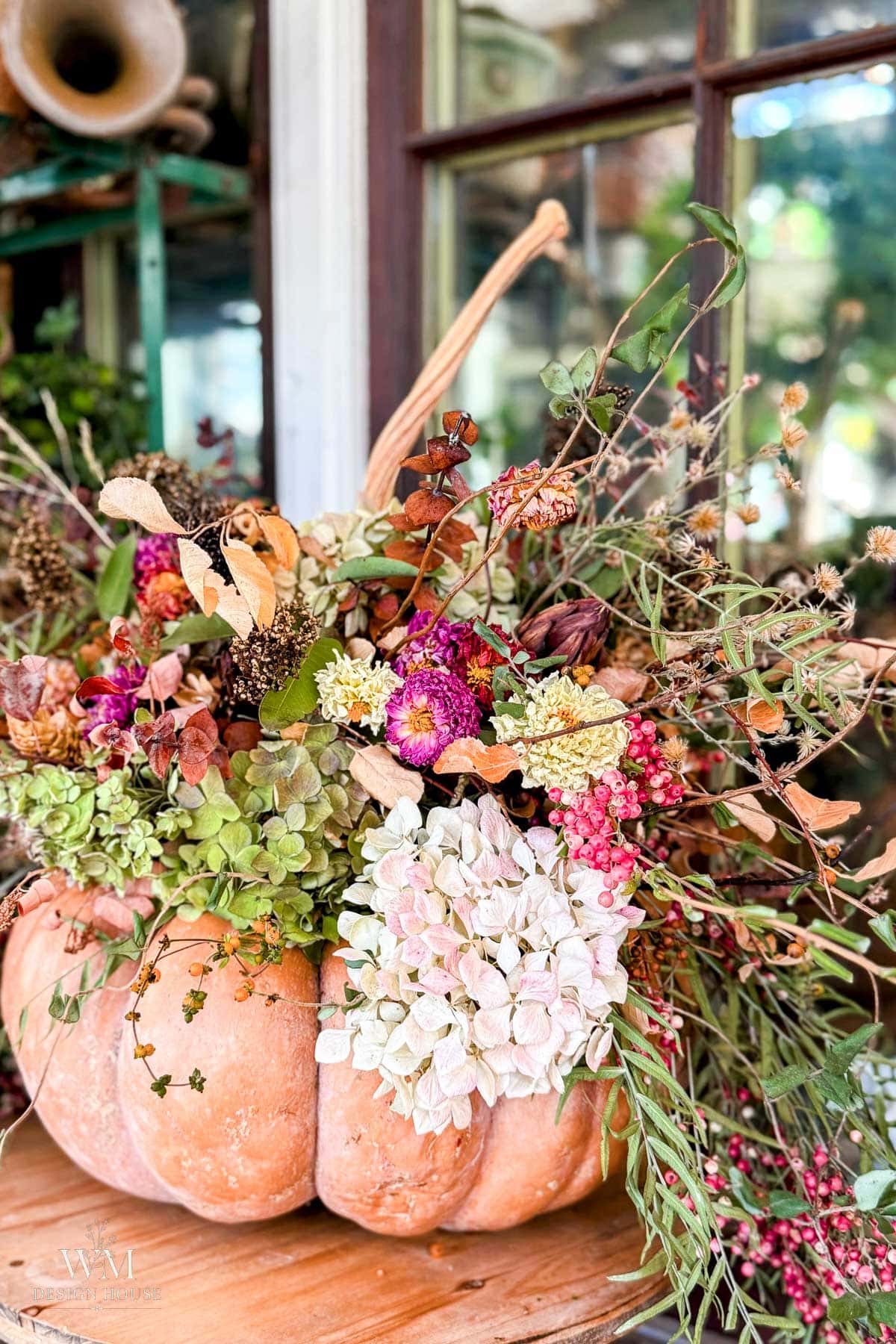 A pumpkin is used as a vase, filled with dried and fresh flowers, greenery, and branches, sitting on a wooden surface in front of a window.