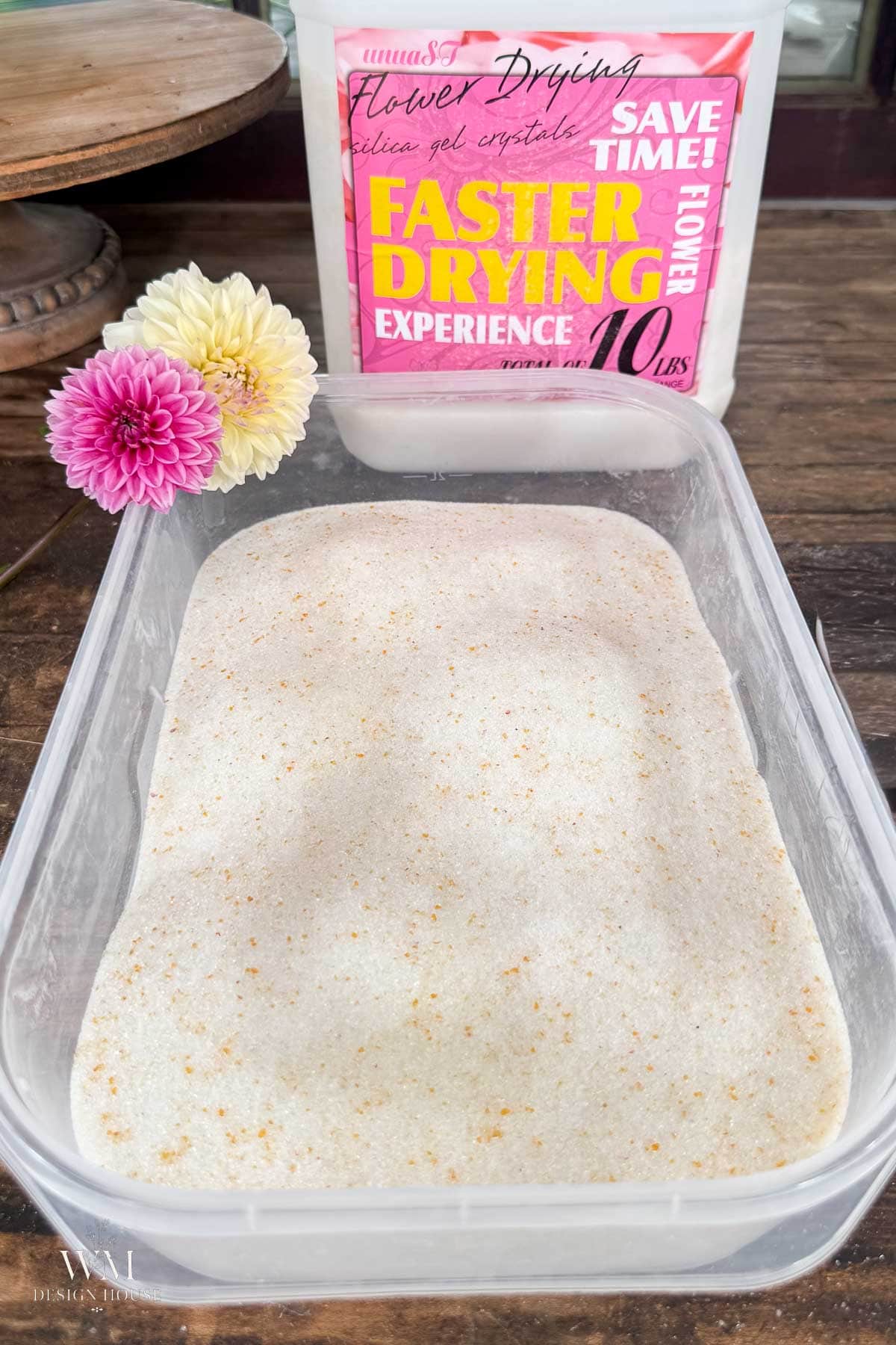 A clear plastic container filled with a white, grainy substance sits on a table. Behind it is a pink and white package labeled “Faster Drying Experience” and two colorful flowers are placed beside the package.