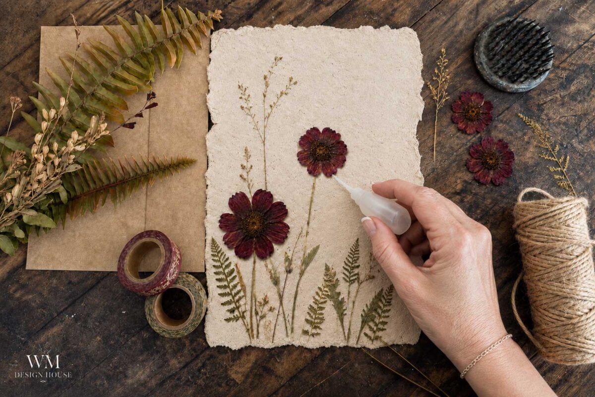 A hand gluing pressed flowers and ferns onto textured paper, surrounded by twine, washi tape, dried plants, and crafting supplies on a wooden table.