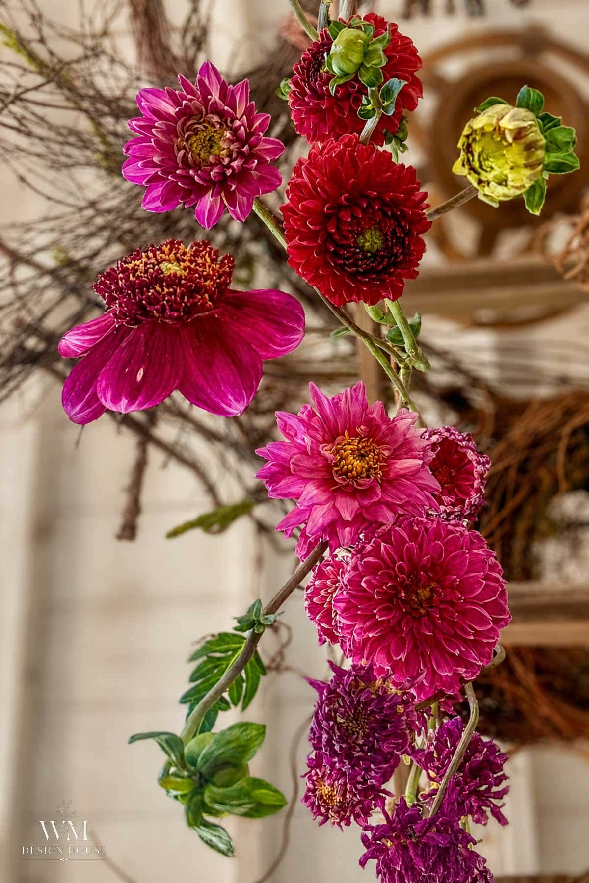 A vibrant arrangement of pink, red, and yellow zinnias with green leaves, set against a blurred background of twigs and a light-colored wall.