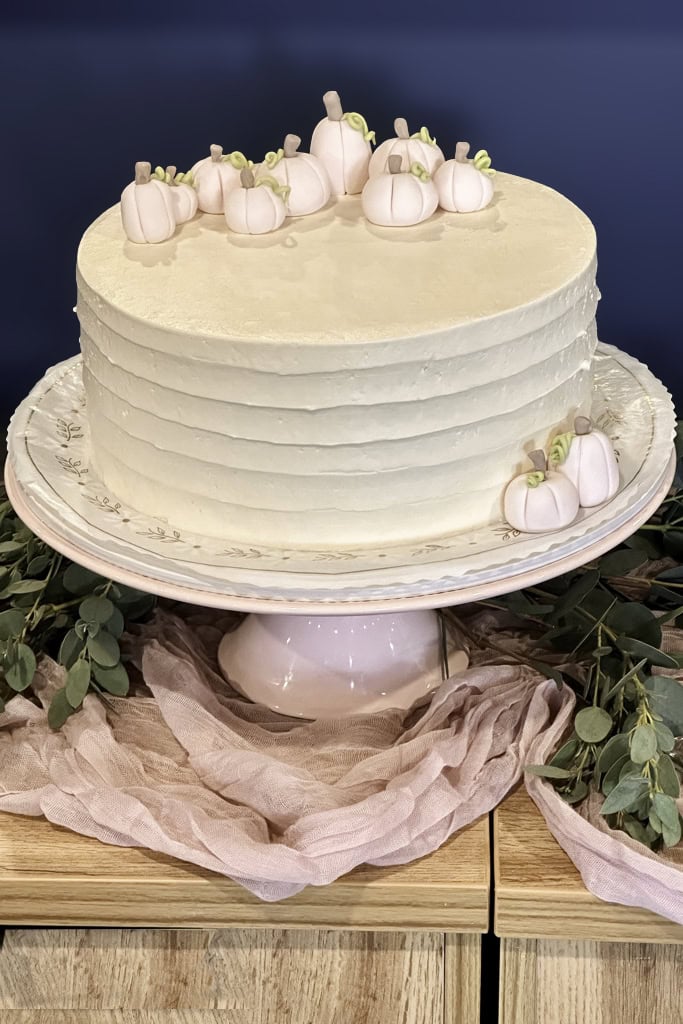 A round white frosted cake topped with small, pale pink pumpkin decorations sits on a white stand, surrounded by greenery and pink fabric.