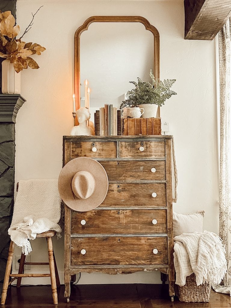 A rustic wooden dresser with white knobs holds books, candles, a plant, and decor. A tan hat hangs on an adjacent chair draped with a blanket. A large mirror and basket with blankets complete the cozy scene.