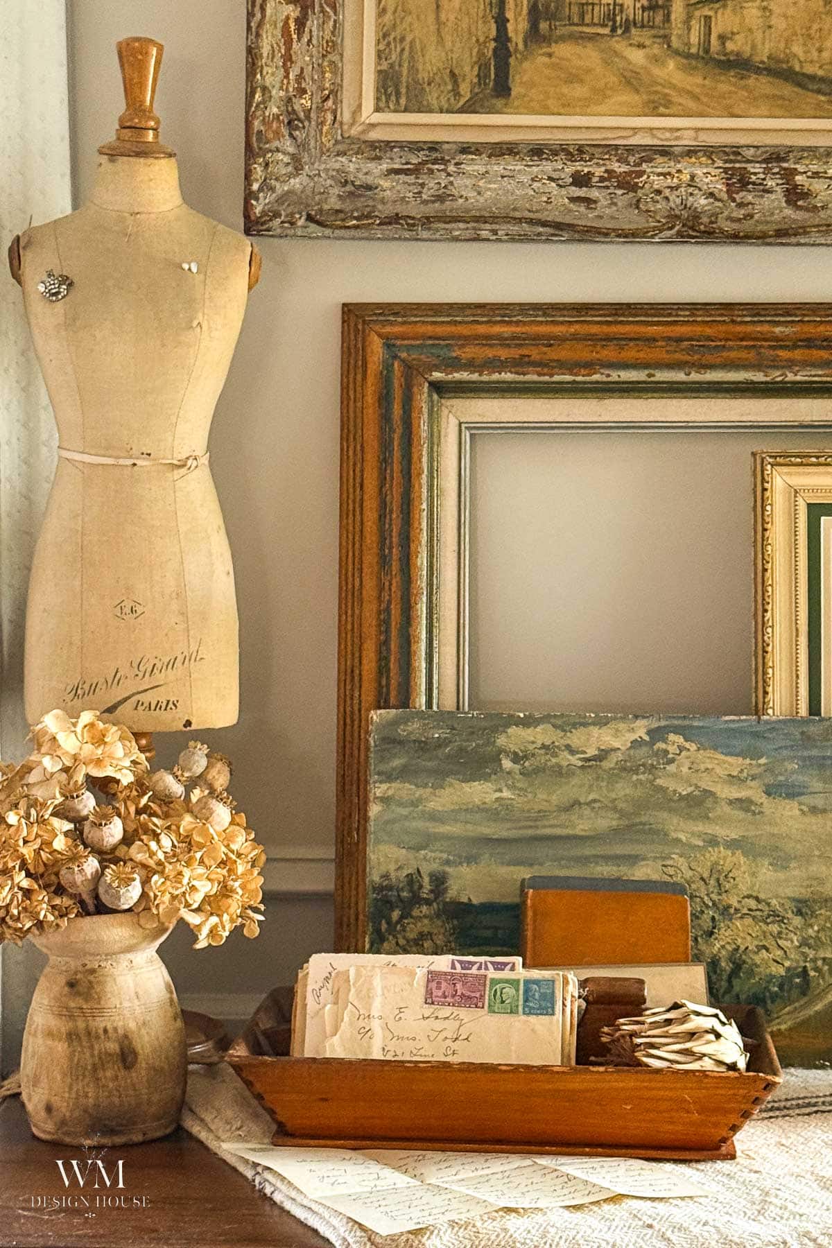 Vintage decor scene with a dress form, dried flowers in a vase, stacked antique frames, a tray holding old letters, postcards, and a small book, all on a lace-covered table.