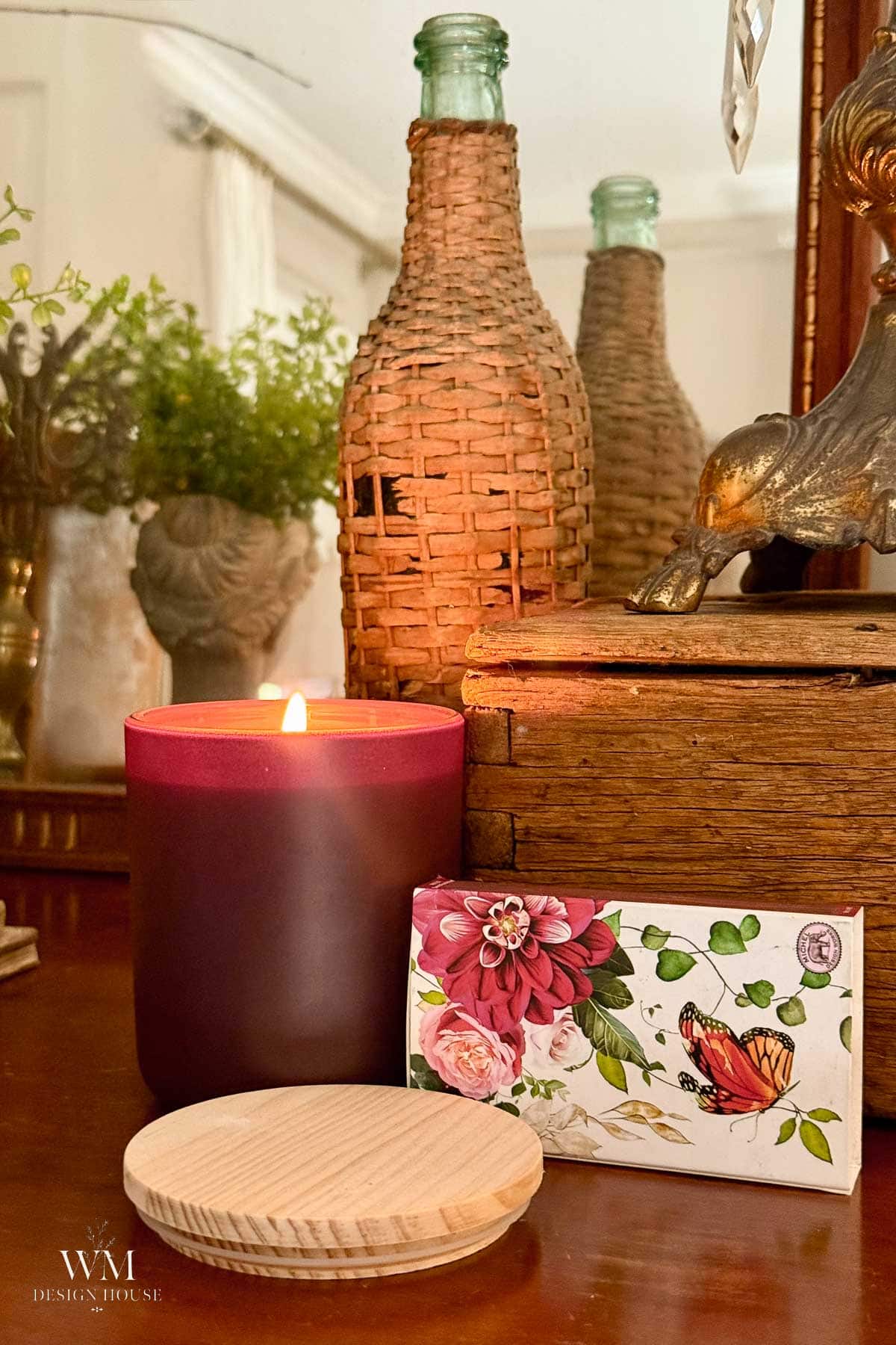 A lit maroon candle, a floral card with a butterfly, a round wooden lid, wicker bottle, and greenery sit on a wooden surface in front of a mirror reflecting the scene.