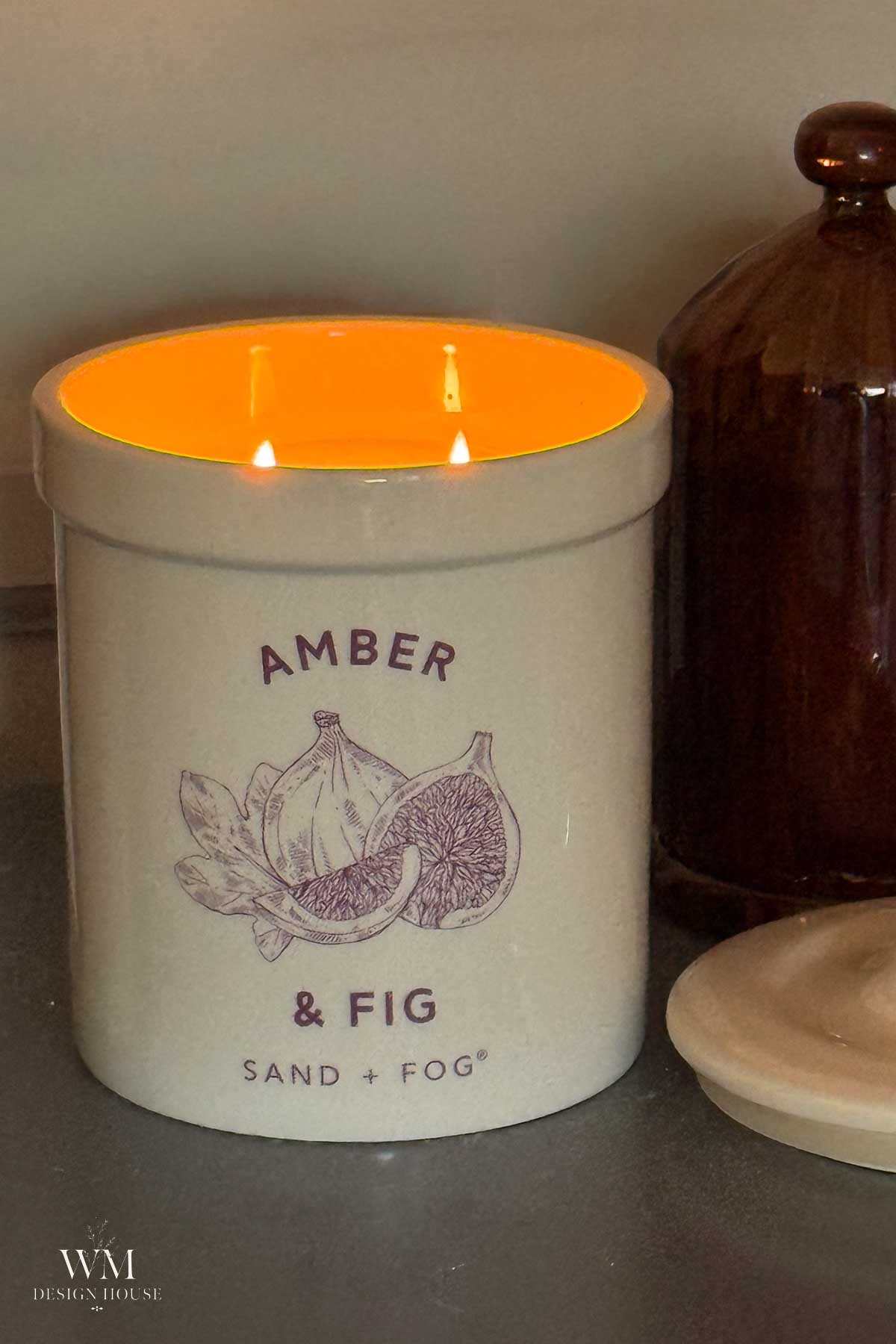 A lit amber and fig scented candle in a cream jar with fig illustrations, next to a brown jar and a lid on a gray surface.