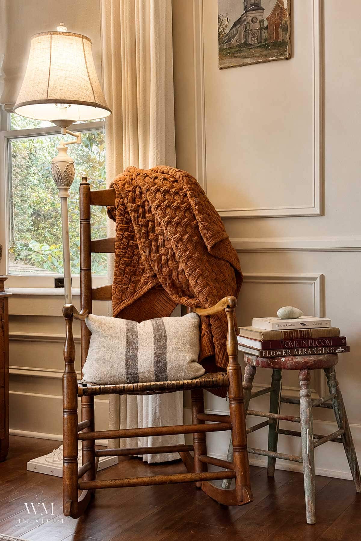 A cozy corner with a wooden rocking chair, rust-colored knitted blanket, striped pillow, small side table with stacked books, and a lamp by a window with beige curtains. Warm, inviting, and vintage decor.