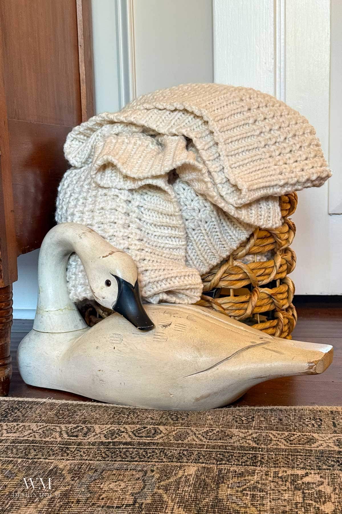 A woven basket with folded chunky cream knit blankets sits beside a wooden swan figurine on a patterned rug, near a wooden cabinet and a white door.