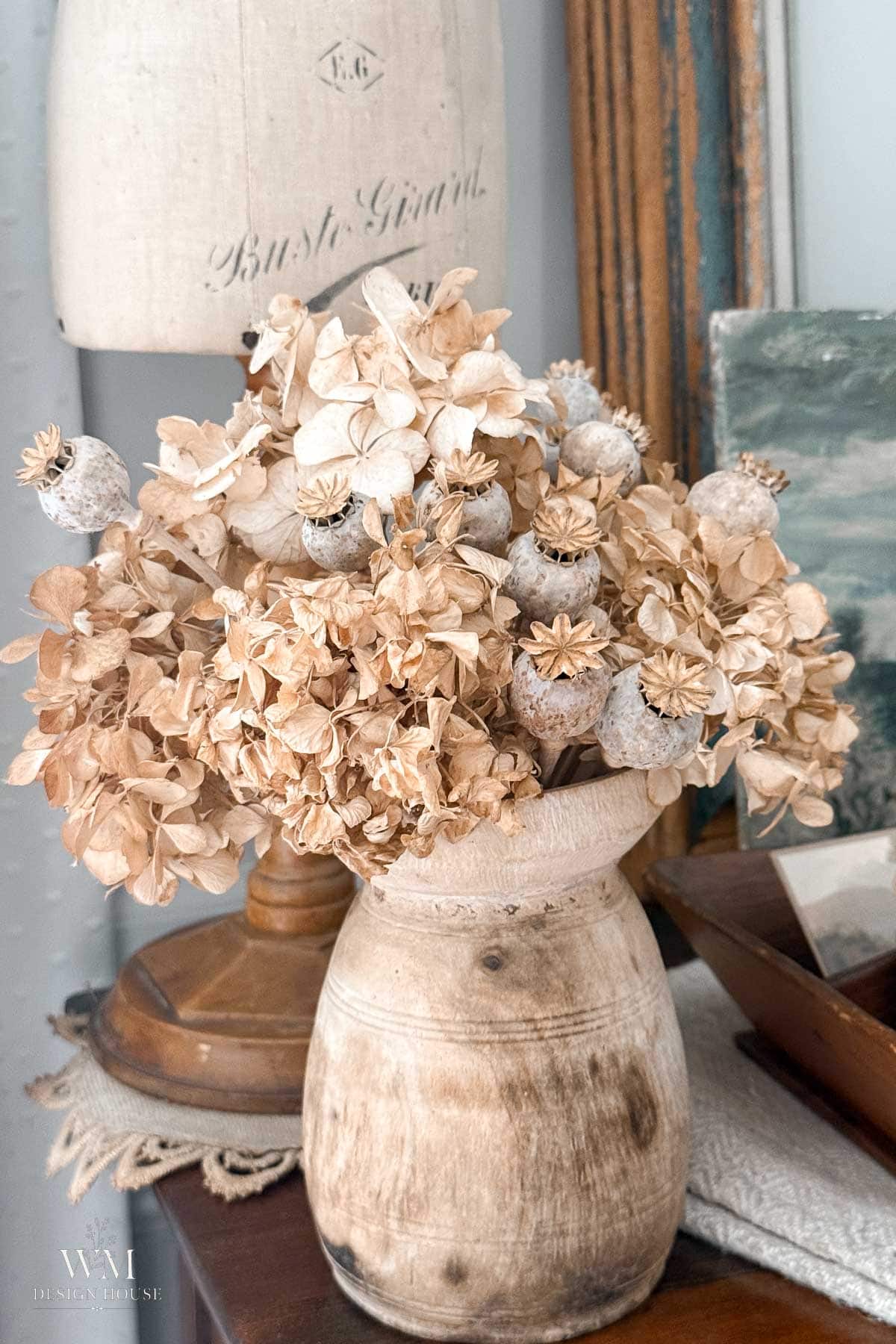A rustic beige vase filled with dried hydrangeas and poppy pods sits on a wooden table, surrounded by vintage decor including books, a lamp, and a mannequin with French script.