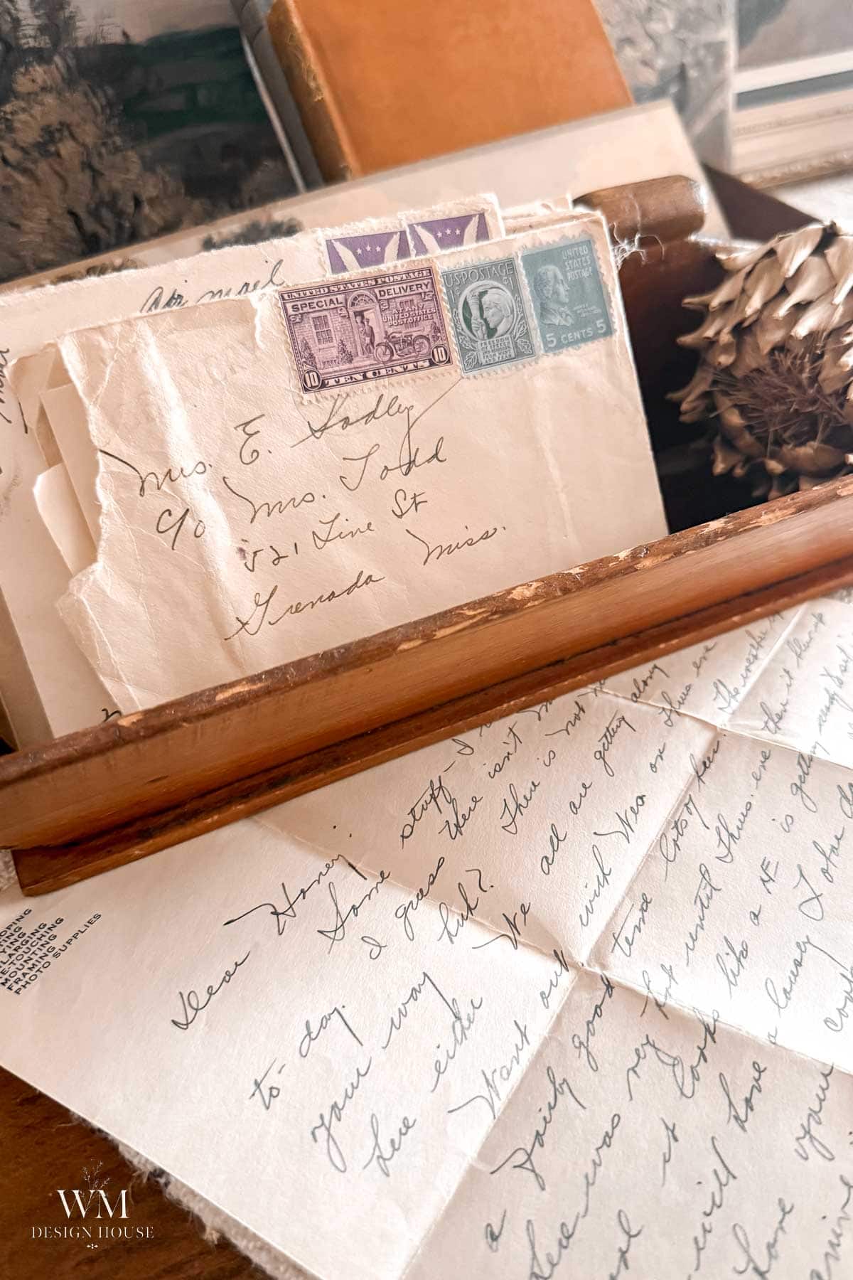 A wooden box holds vintage letters with postage stamps. An unfolded handwritten letter lies in front, and a dried flower rests nearby. Old books and sepia-toned photos are in the background.