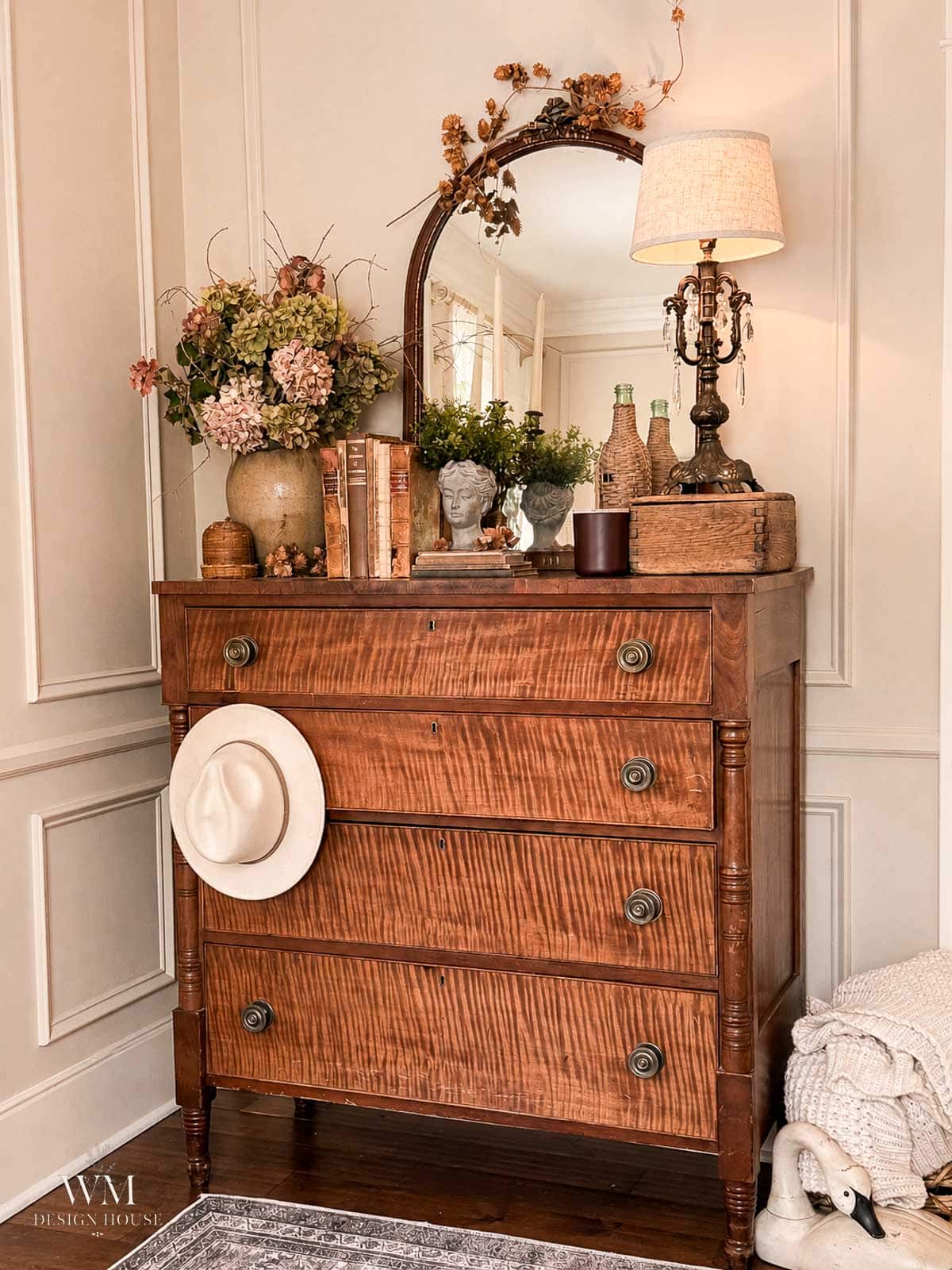 A vintage wooden dresser holds a table lamp, books, vases, dried flowers, and a round mirror. A white hat hangs on a drawer, and a woven basket with blankets sits on the floor beside it.