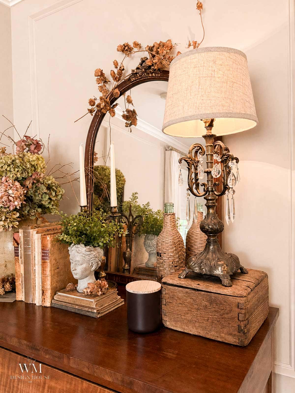 A vintage lamp with a beige shade sits on stacked books, alongside a mirror, dried flowers, old books, a small bust planter with greenery, and decorative glass bottles on a wooden dresser.