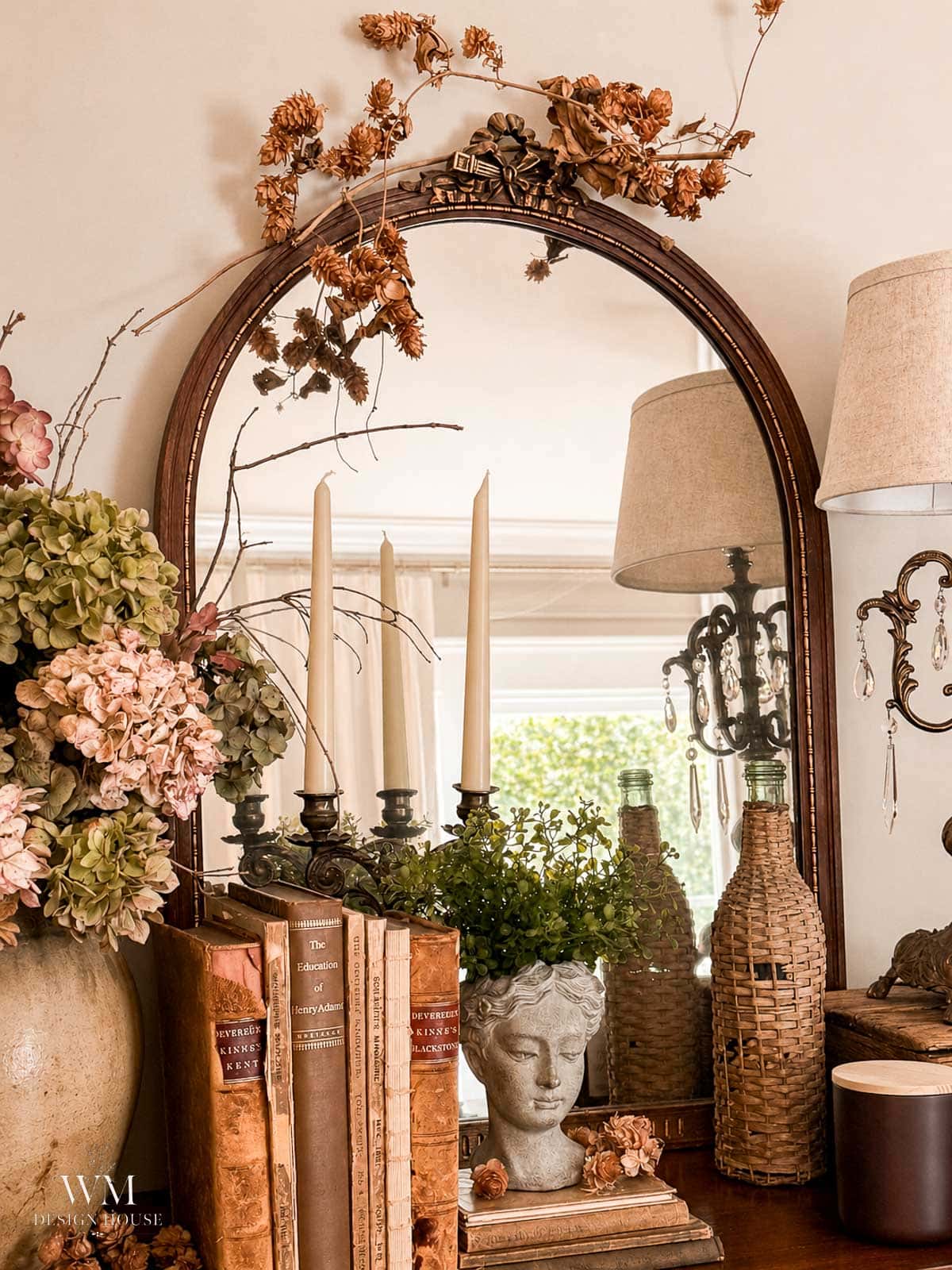 A vintage mirror reflects candlesticks, books, a bust planter with greenery, woven bottles, and dried flowers, creating a cozy, antique-inspired vignette on a wooden table.
