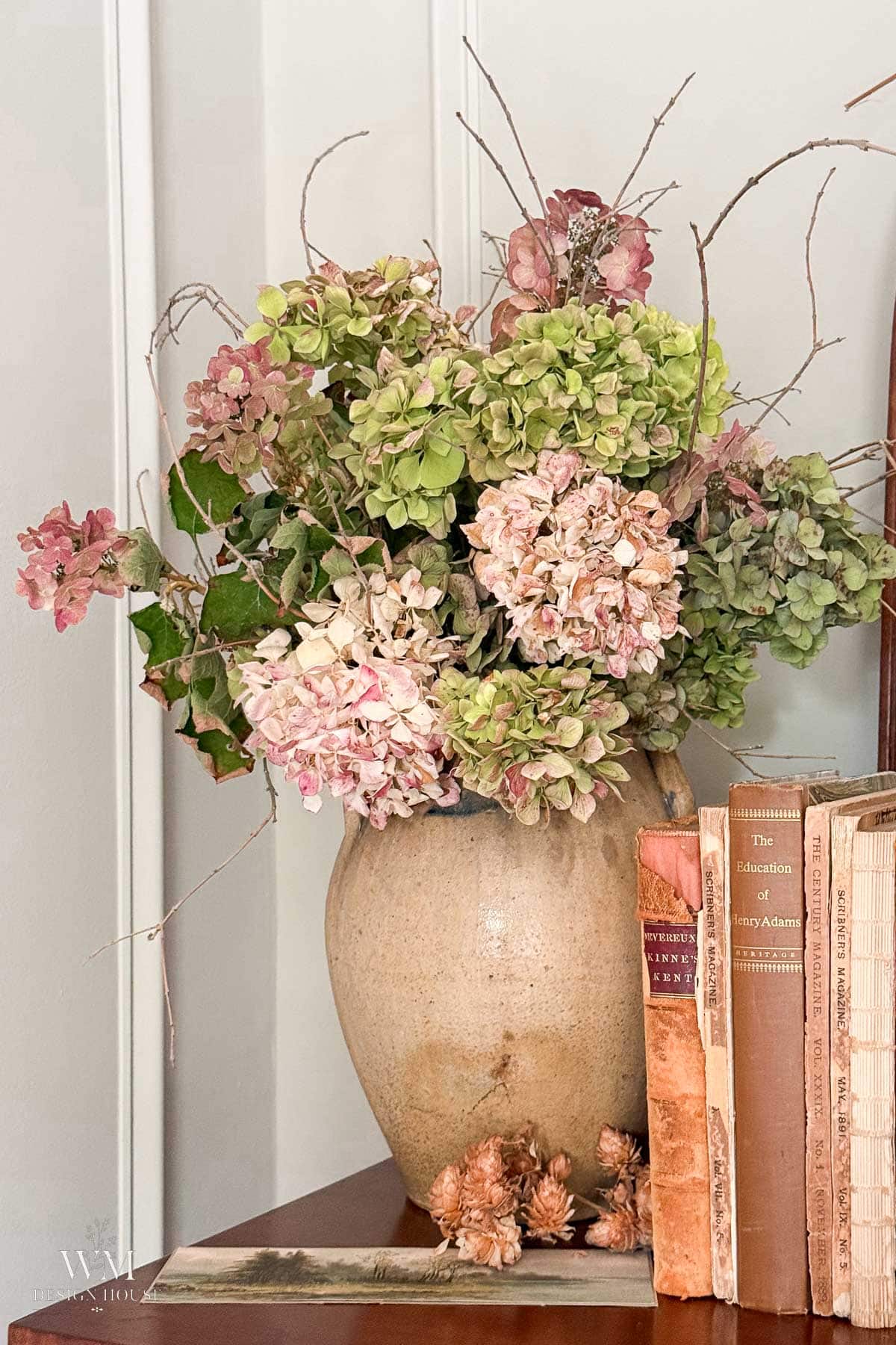 A ceramic vase with pink and green dried hydrangeas sits on a wooden surface next to a row of vintage books and a small cluster of pinecones.