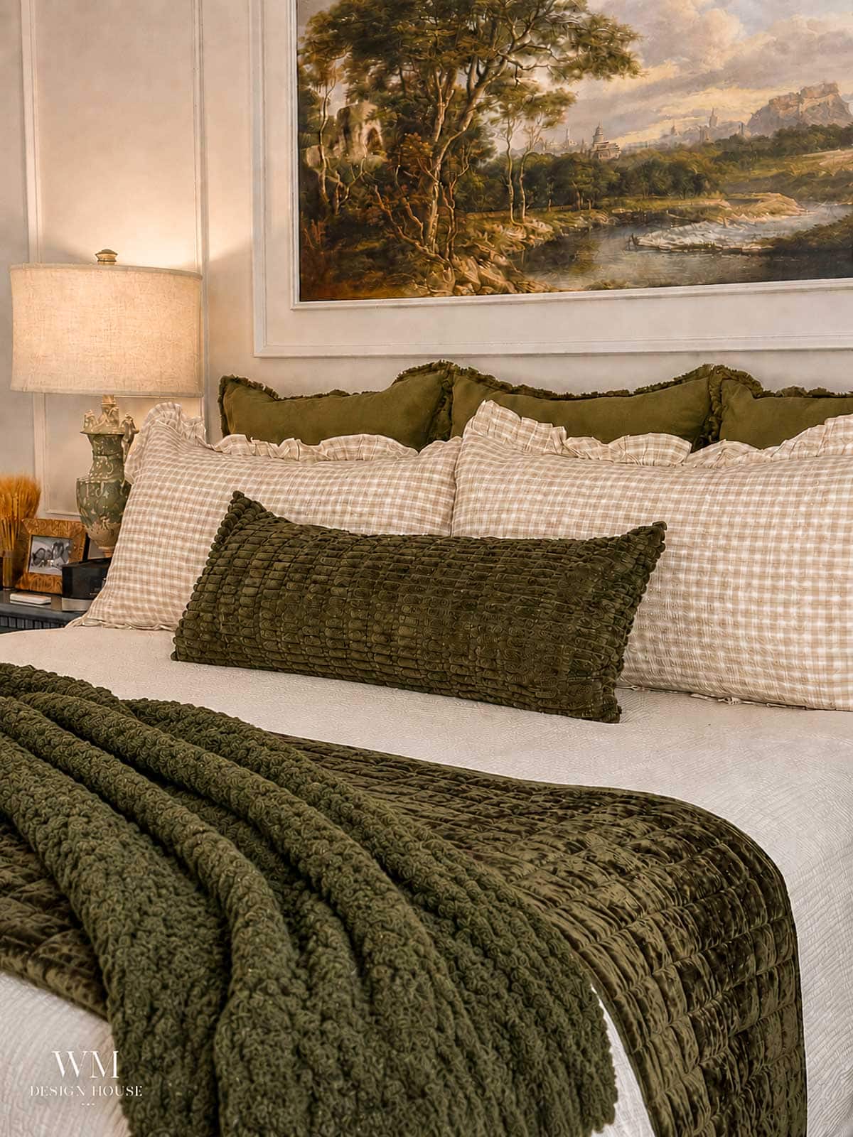 A neatly made bed with green and cream checked pillows, a textured green pillow, and a green blanket. A table lamp and a landscape painting hang above the bed. The room has a cozy and inviting feel.