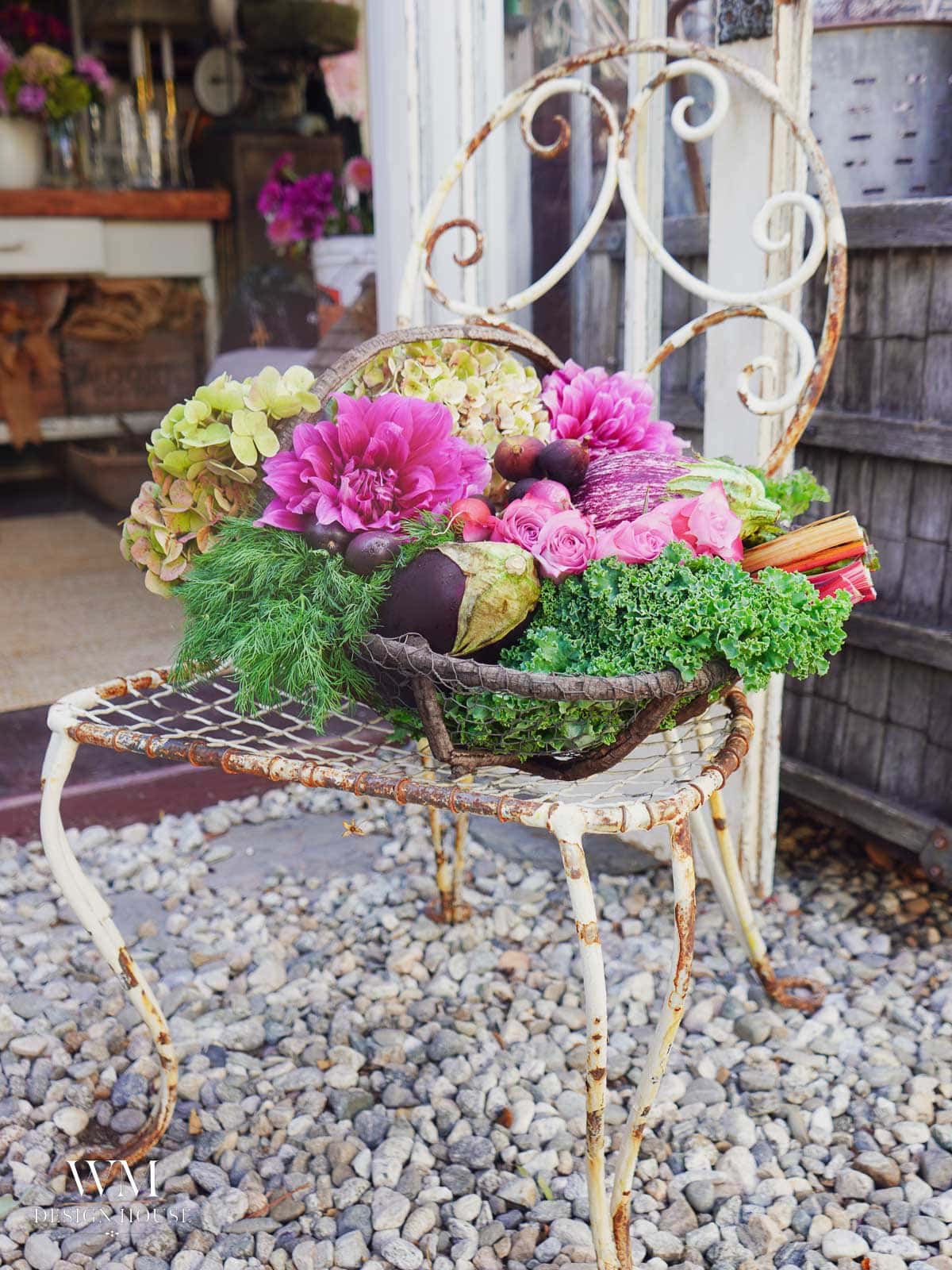 A rustic metal chair holds a basket filled with pink flowers, greenery, and vegetables, sitting on a gravel surface outside a garden shop.