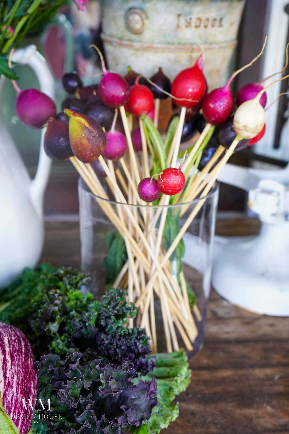 A clear glass holds colorful radishes on wooden skewers, arranged like a bouquet. Fresh kale and other leafy greens sit on a rustic wooden table nearby.