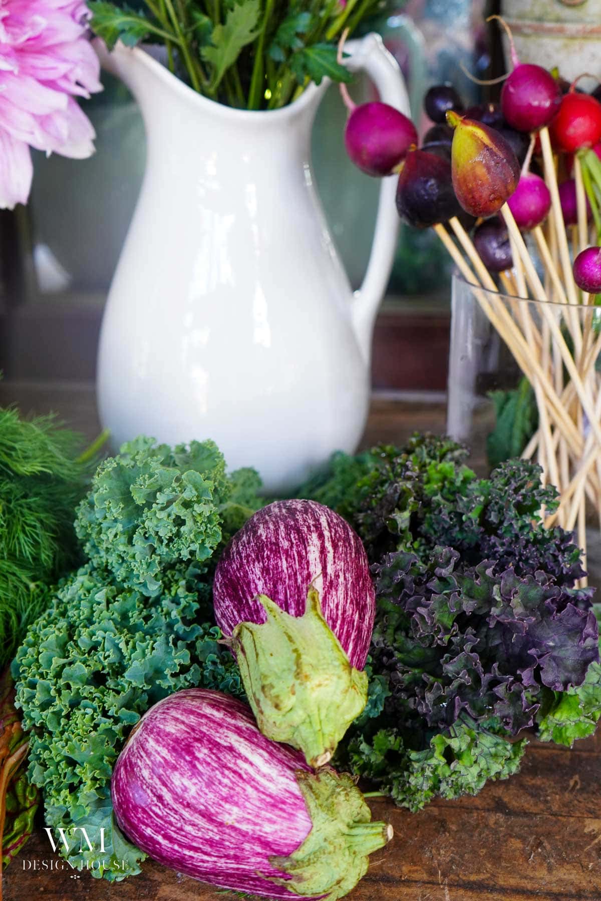 Two striped eggplants and bunches of curly kale are arranged on a wooden surface, with a white pitcher and a glass holding skewered radishes and flowers in the background.
