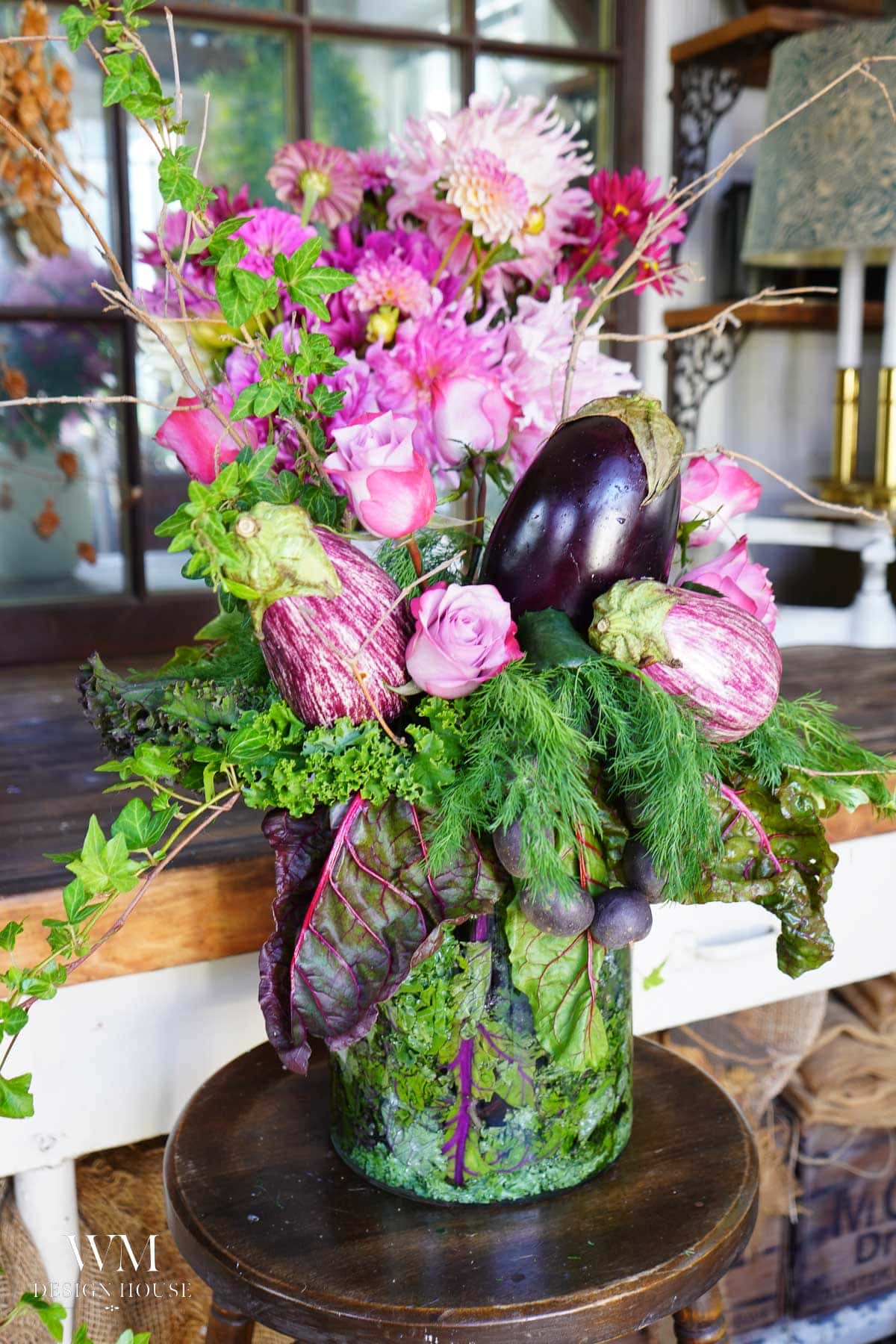 A vibrant floral arrangement in a glass vase features pink flowers, leafy greens, and two eggplants. The bouquet is set on a wooden table with a window and decor in the background.