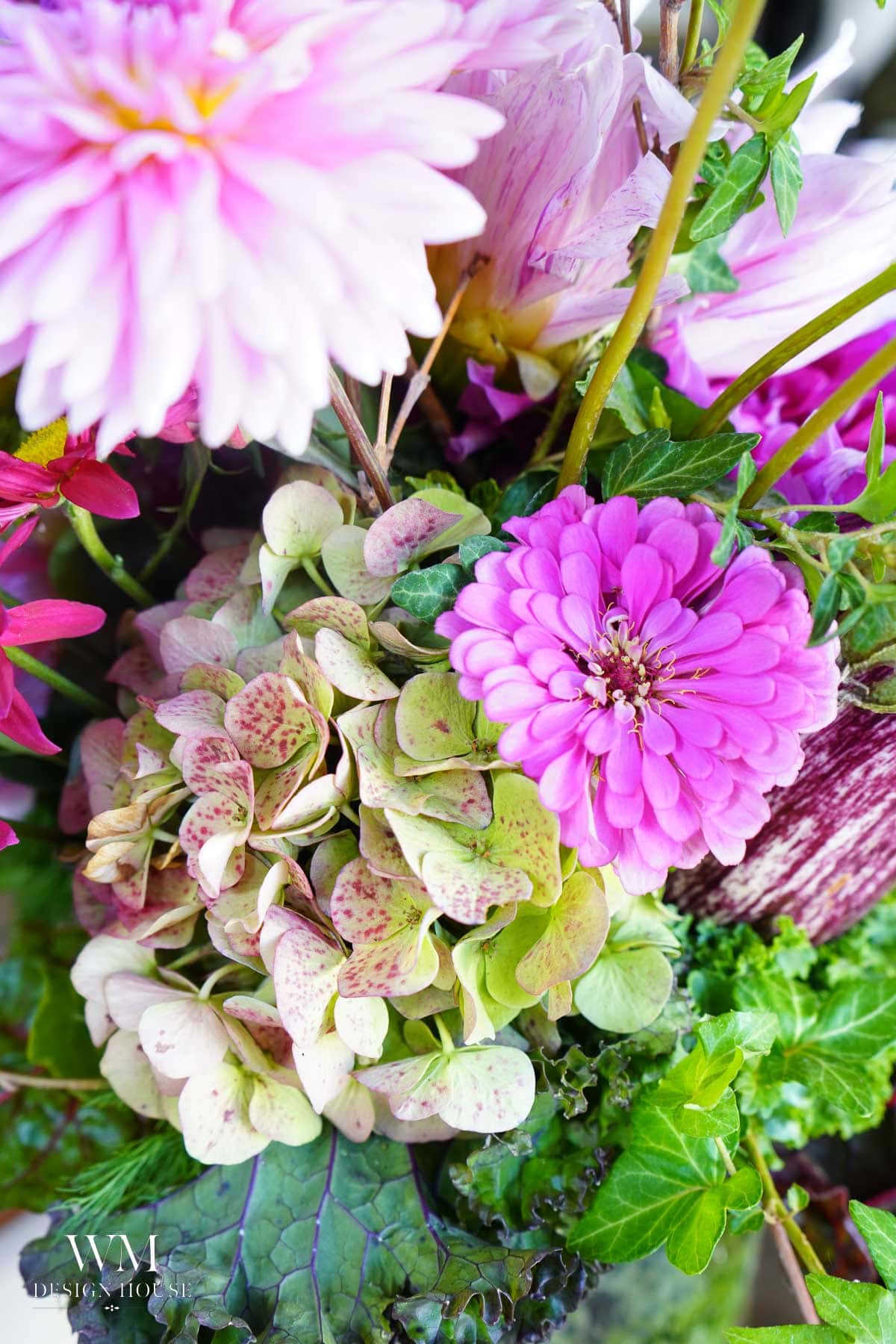 A close-up of a vibrant floral arrangement with pink zinnias, pale-green hydrangeas with red speckles, and various green leaves. The flowers appear fresh and colorful.