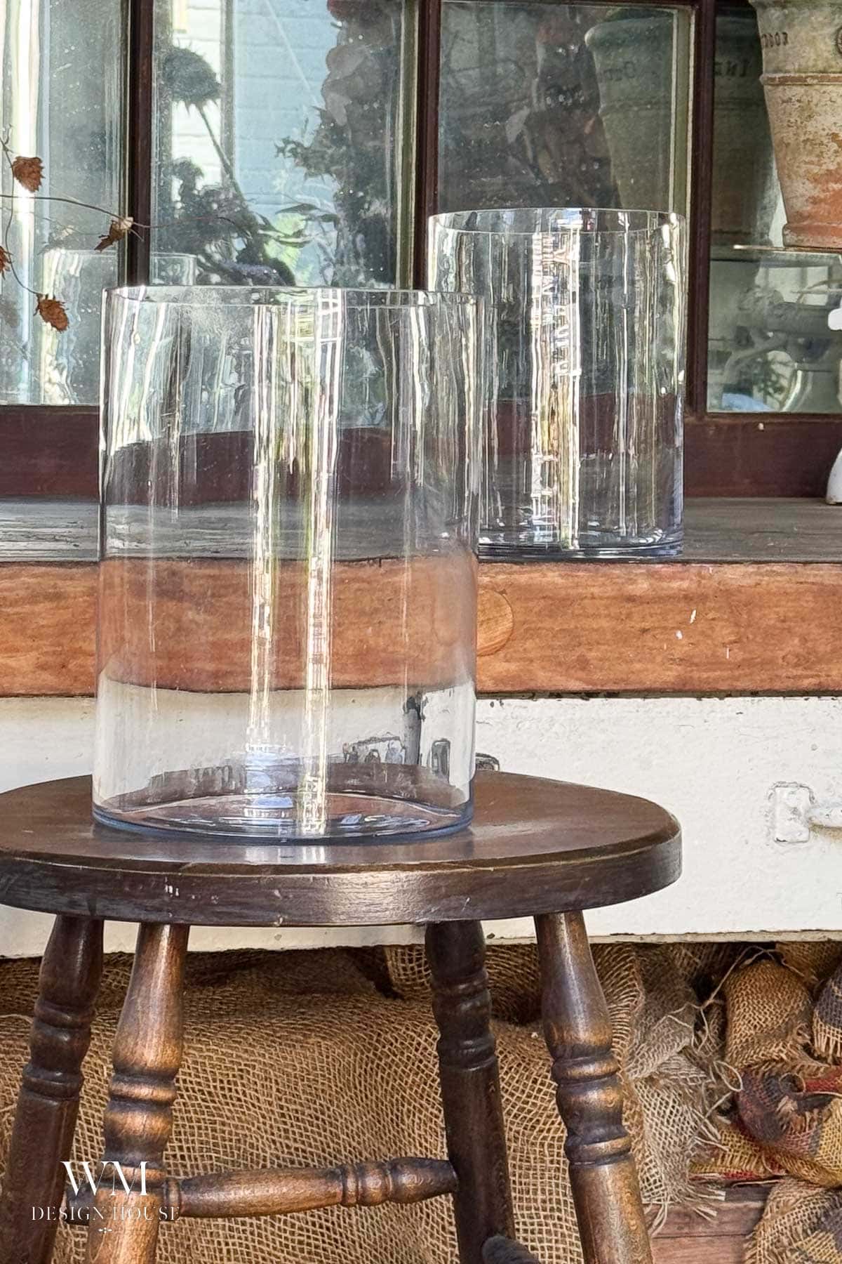 Two clear glass vases sit, one on a wooden stool in the foreground and another on a wooden surface behind it. A rustic window, burlap sacks, and some potted plants appear in the background.