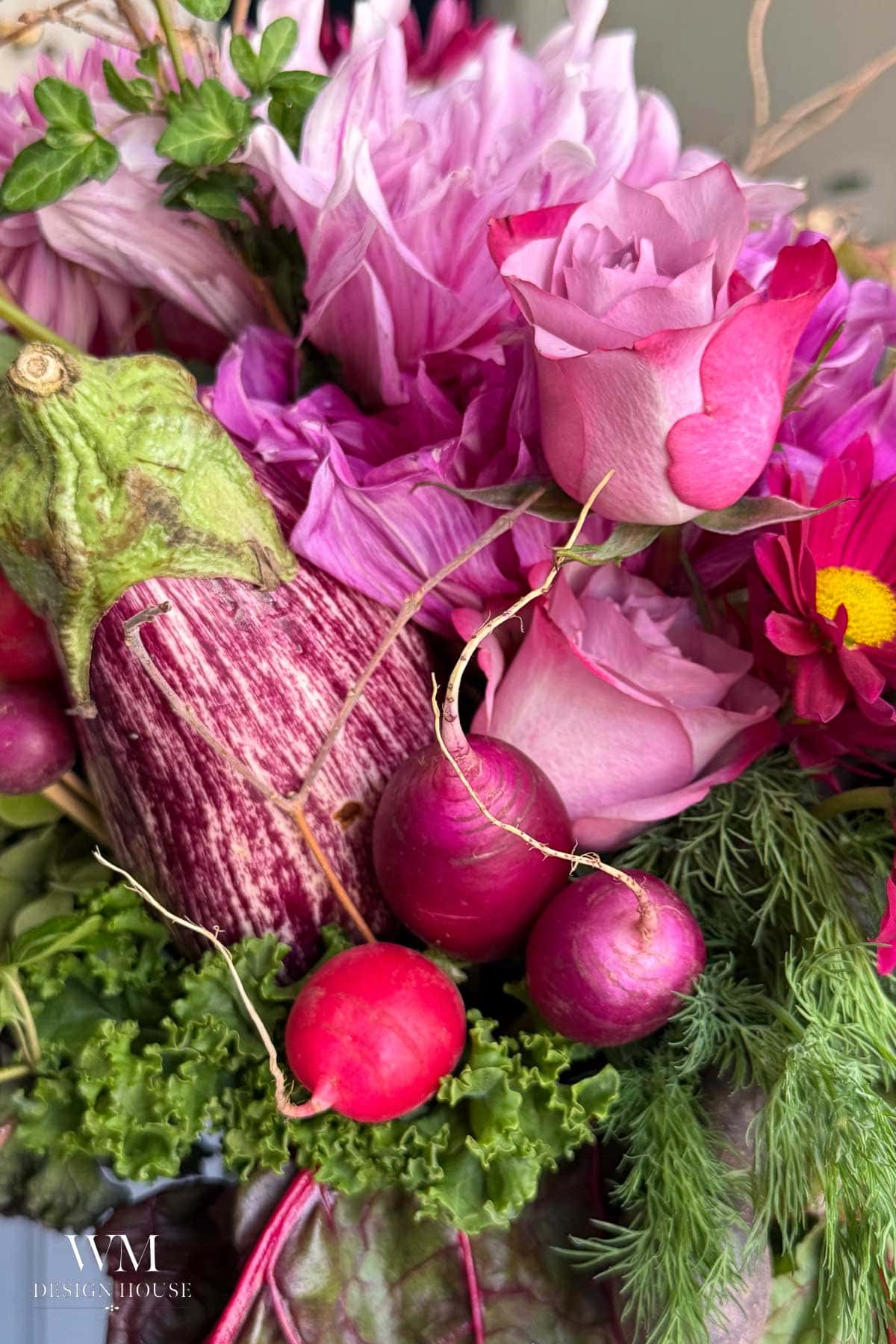 A vibrant floral arrangement featuring pink roses, striped eggplant, radishes, and assorted greenery, blending fresh vegetables and flowers in shades of pink, purple, and green.