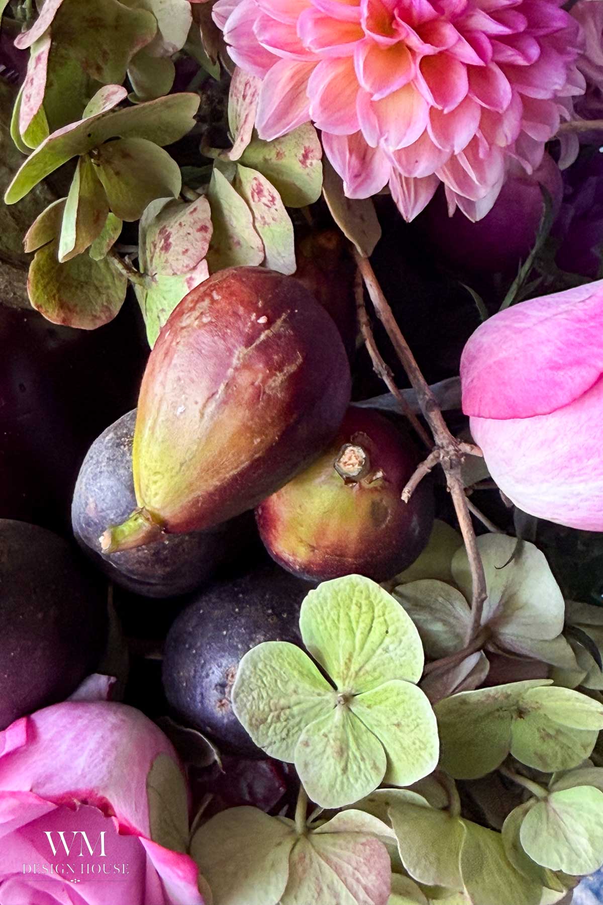 Close-up of fresh figs, purple plums, and hydrangea flowers with pink dahlias, creating a colorful, textured arrangement. The WM Design House logo appears in the bottom left corner.