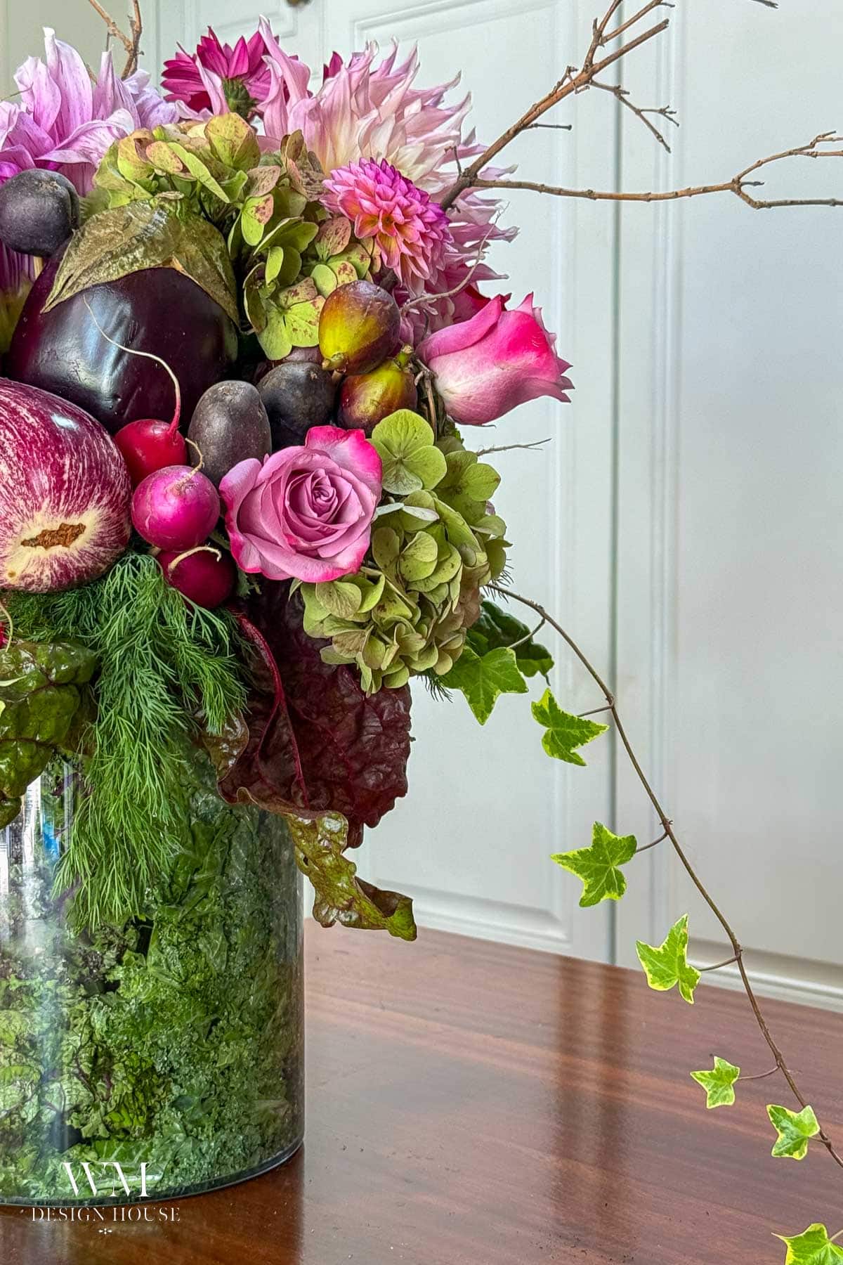 A floral arrangement in a glass vase with roses, dahlias, greenery, vegetables like eggplant and radishes, and ivy trailing out. The arrangement sits on a wooden table against a white background.