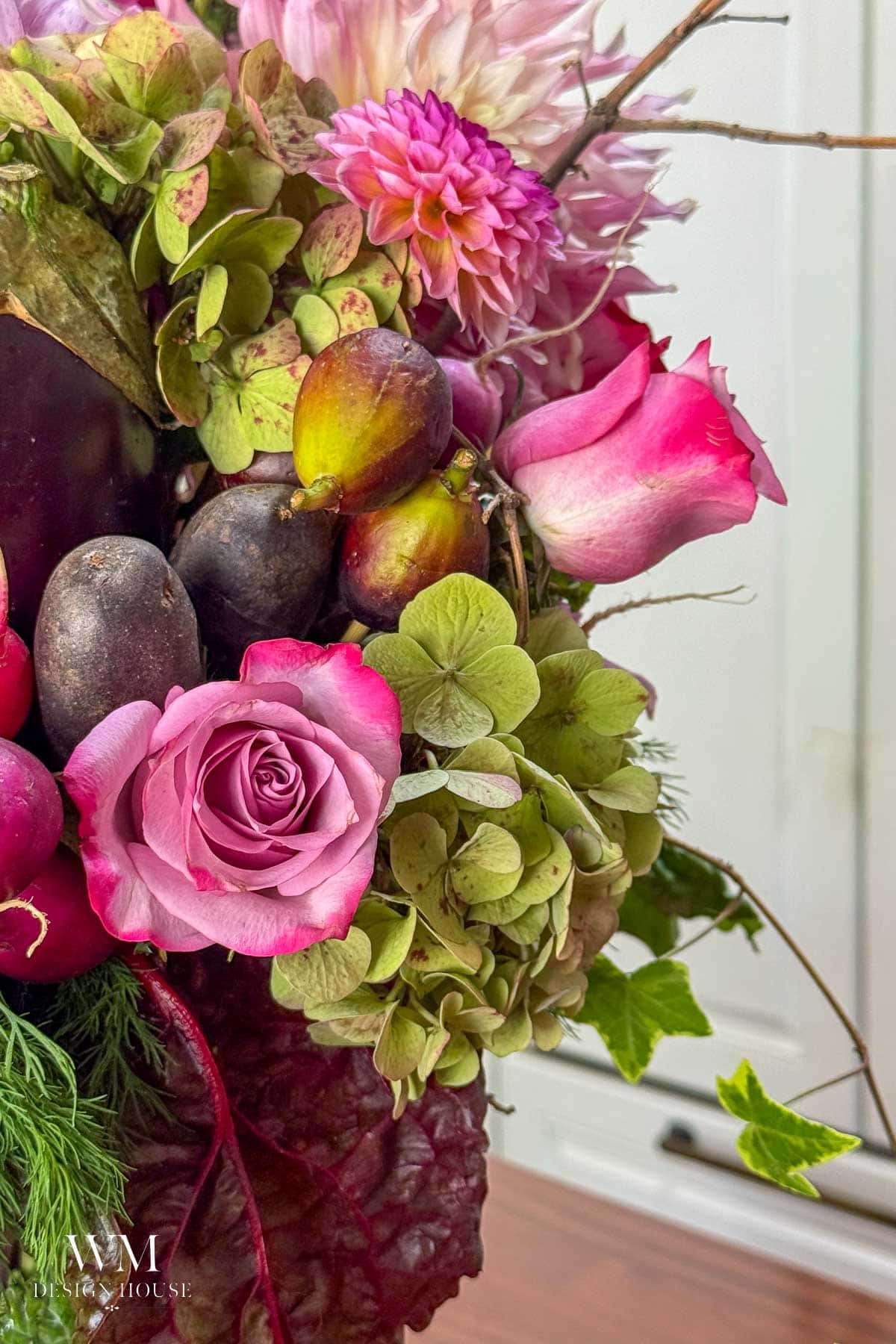 A close-up of a vibrant floral arrangement with pink roses, dahlias, green hydrangeas, figs, and dark grapes, set against a light indoor background.