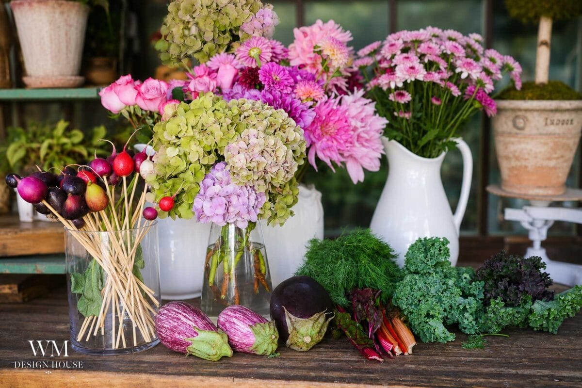 A table displays bouquets of flowers in glass and ceramic vases, with vegetables including eggplants, radishes, kale, and Swiss chard arranged in front, creating a colorful, rustic setting.