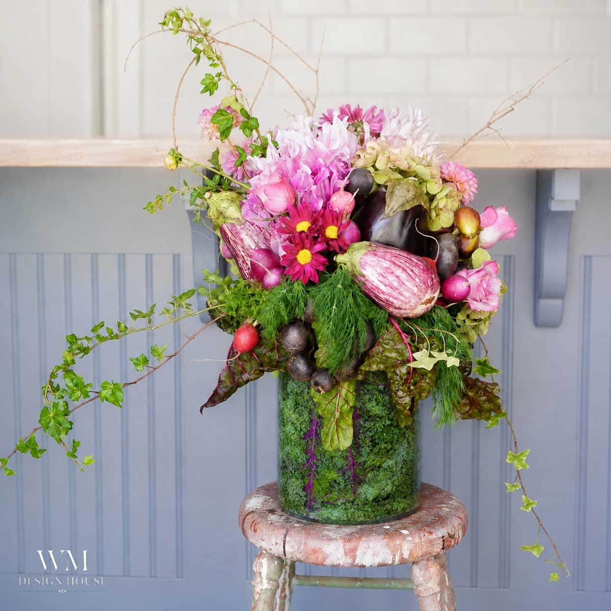 A lush floral arrangement in a glass vase features pink flowers, green foliage, and decorative gourds, displayed on a rustic stool in front of a gray counter and white tile background.