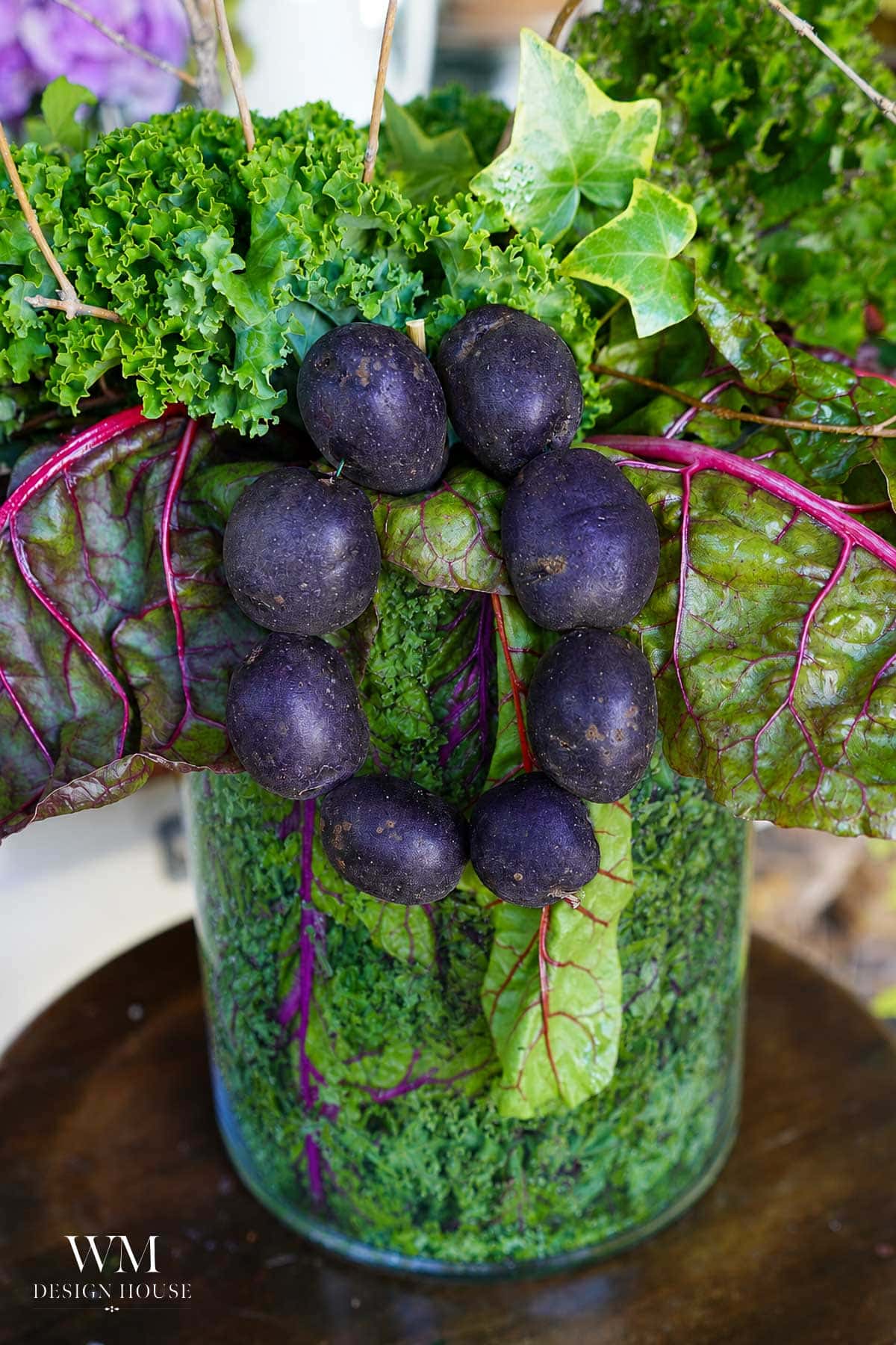 A glass vase filled with green leafy vegetables is decorated with dark purple small potatoes arranged in a circle, and large purple-veined leaves extend outward.