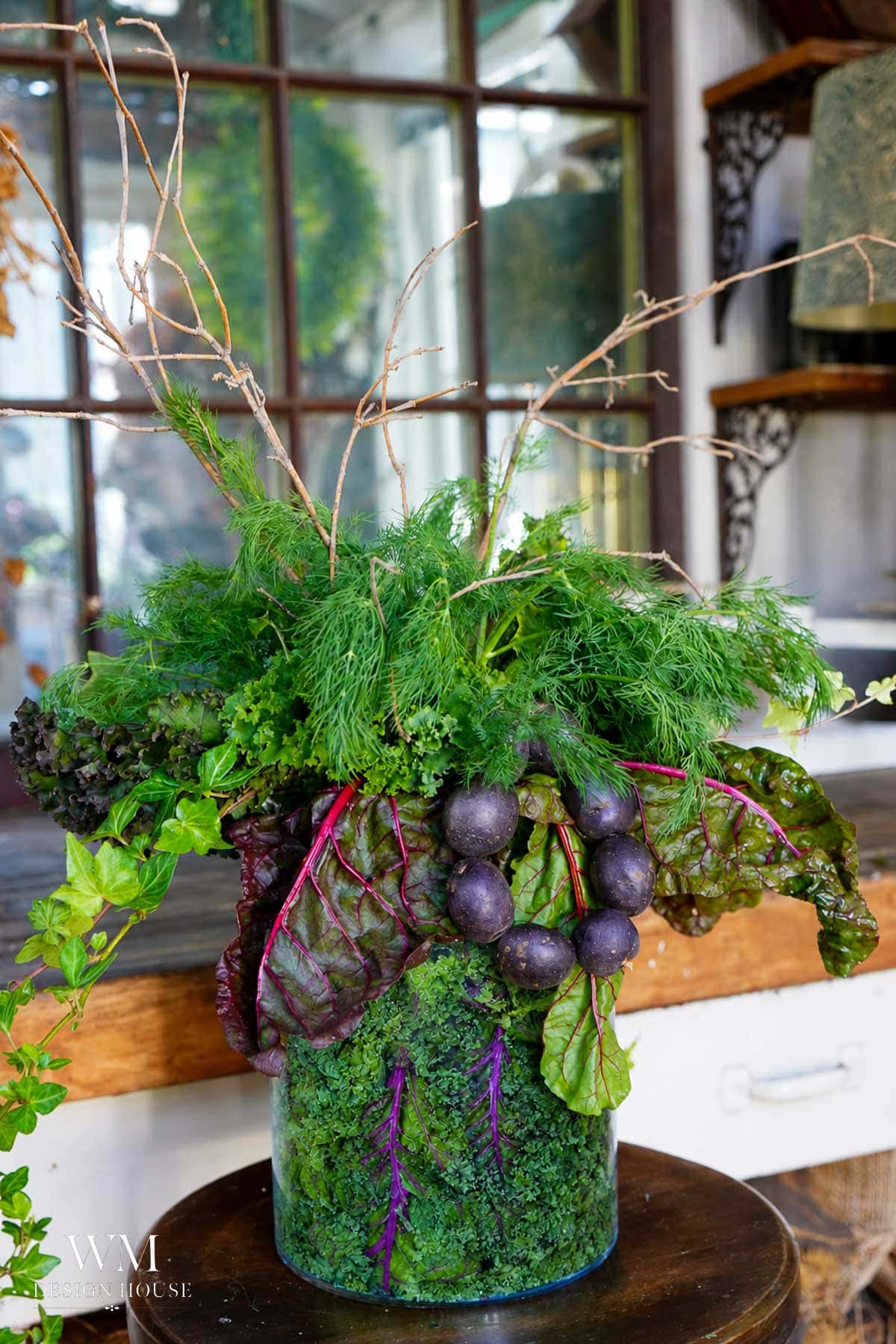 A vibrant arrangement of leafy greens, herbs, and purple radishes in a vase, displayed indoors with a rustic window and shelves in the background. The vase is decorated with greenery and purple stems.