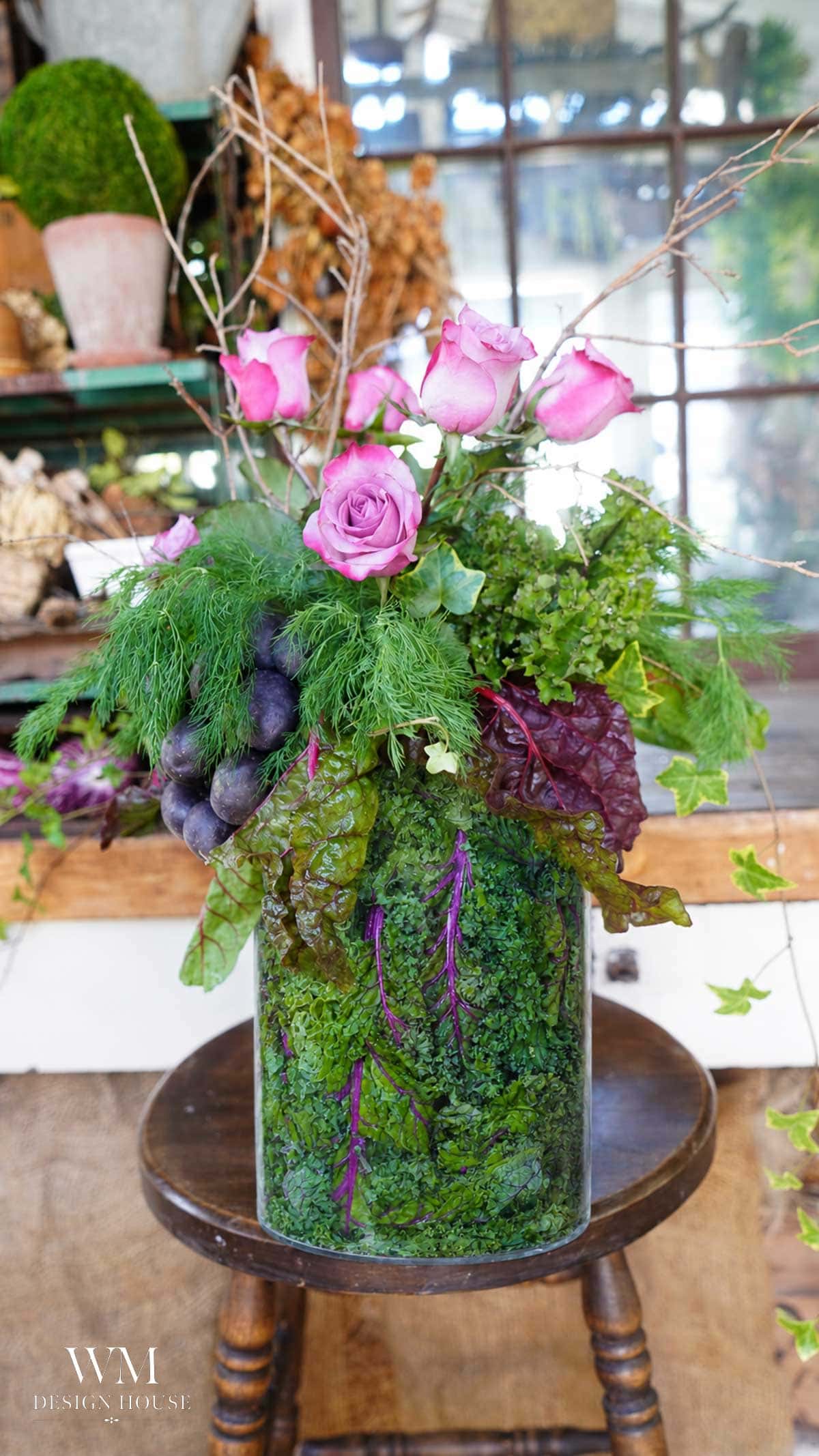 A glass vase filled with moss and greens holds pink roses, leafy greens, grapes, and branches, creating a vibrant, garden-inspired floral arrangement on a wooden chair.