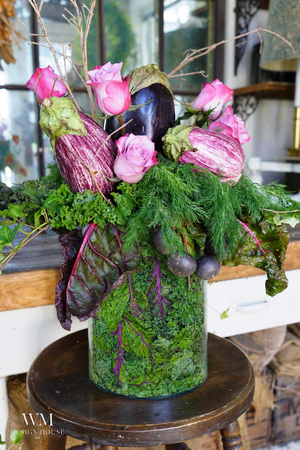 A floral arrangement with pink roses, striped eggplants, leafy greens, and herbs in a green textured vase on a wooden stool indoors.