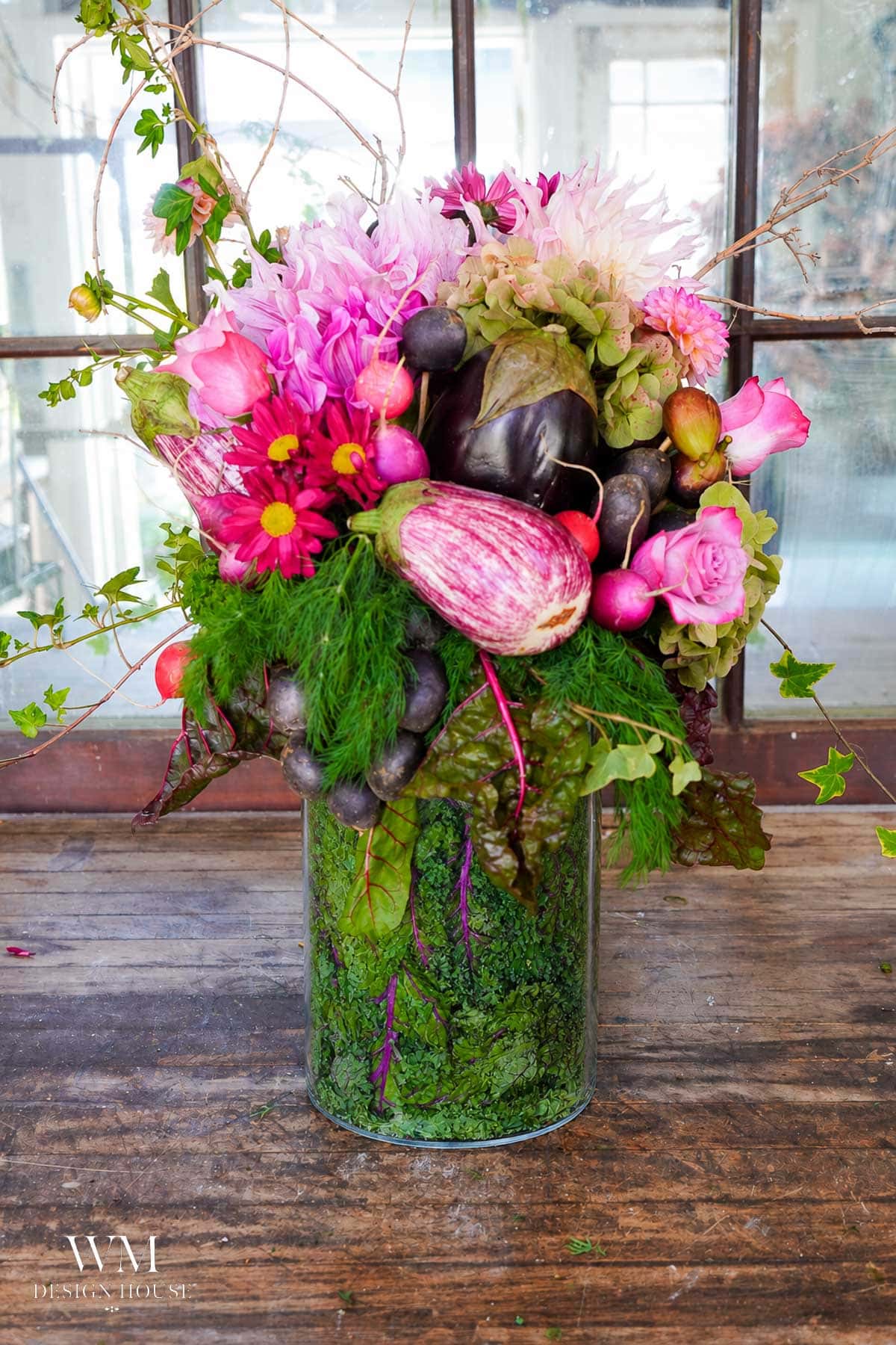 A tall glass vase holds a vibrant arrangement of pink flowers, green leaves, and assorted vegetables including eggplants and radishes, set on a wooden table in front of a window.