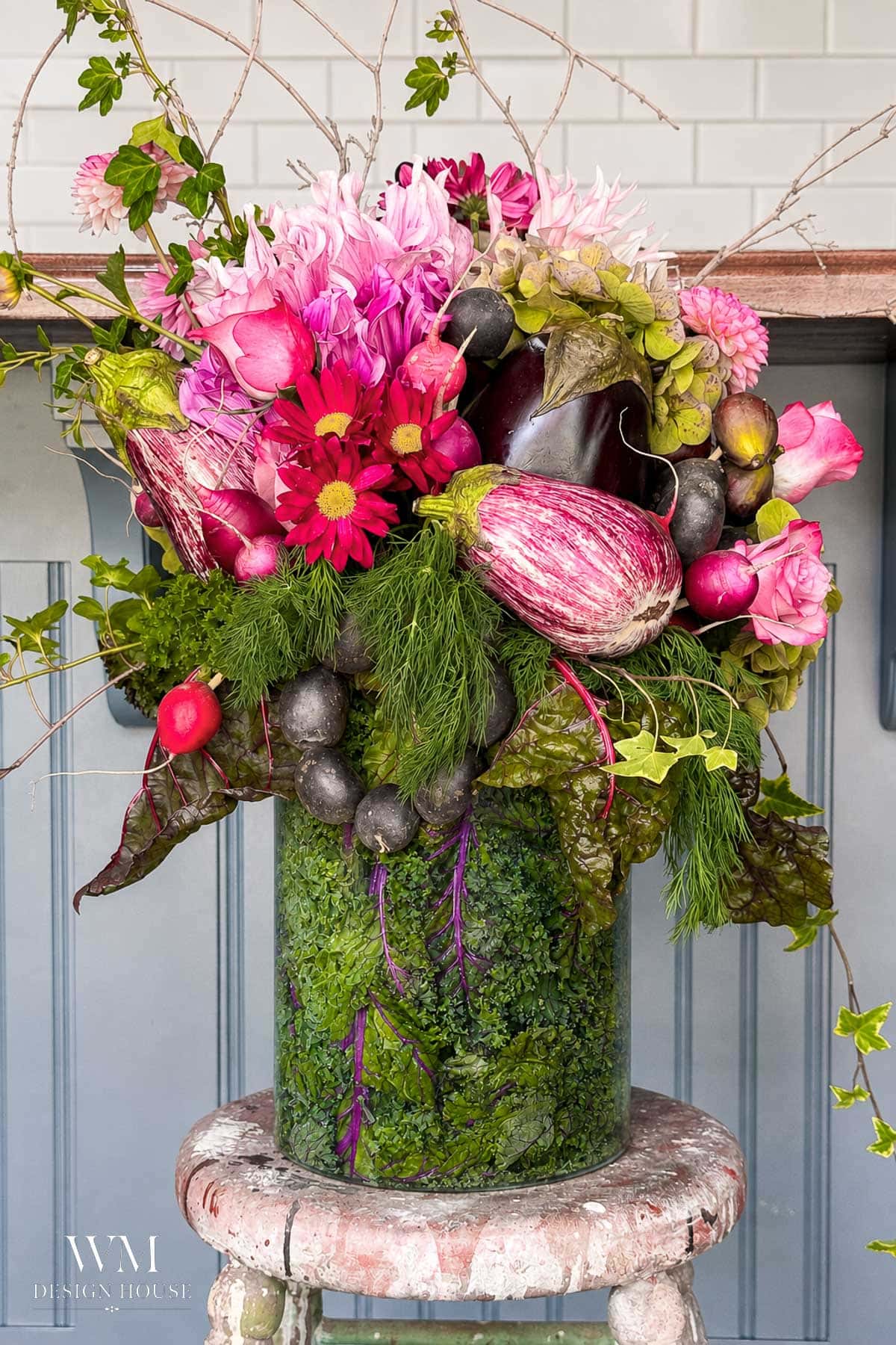 A colorful floral arrangement in a glass vase features pink flowers, leafy greens, striped eggplants, radishes, and herbs, displayed on a weathered round stool against a light blue paneled wall.