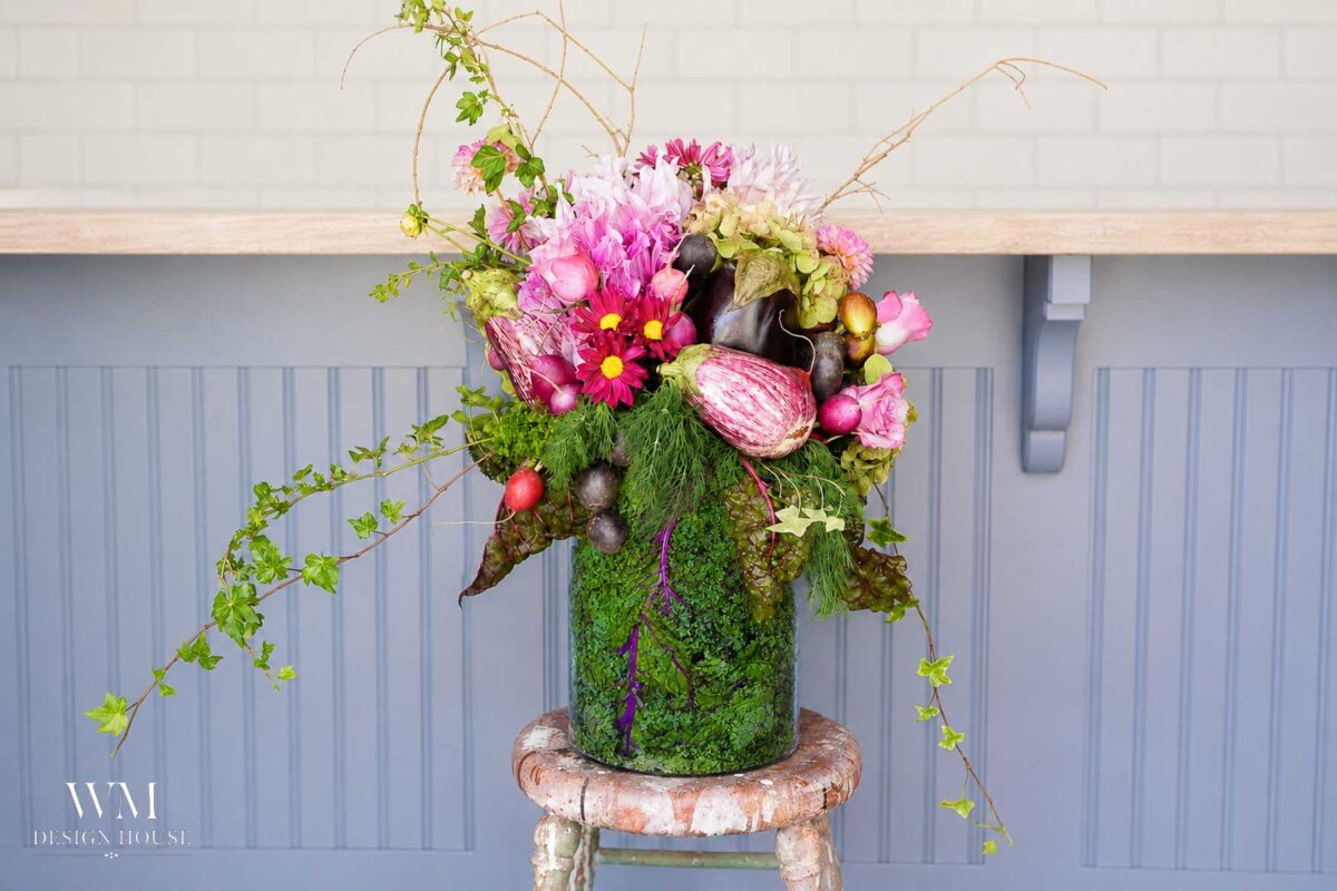 A vibrant floral arrangement in a moss-covered vase, featuring pink and purple flowers, vegetables, and trailing greenery, sits on a rustic wooden stool against a gray paneled background.