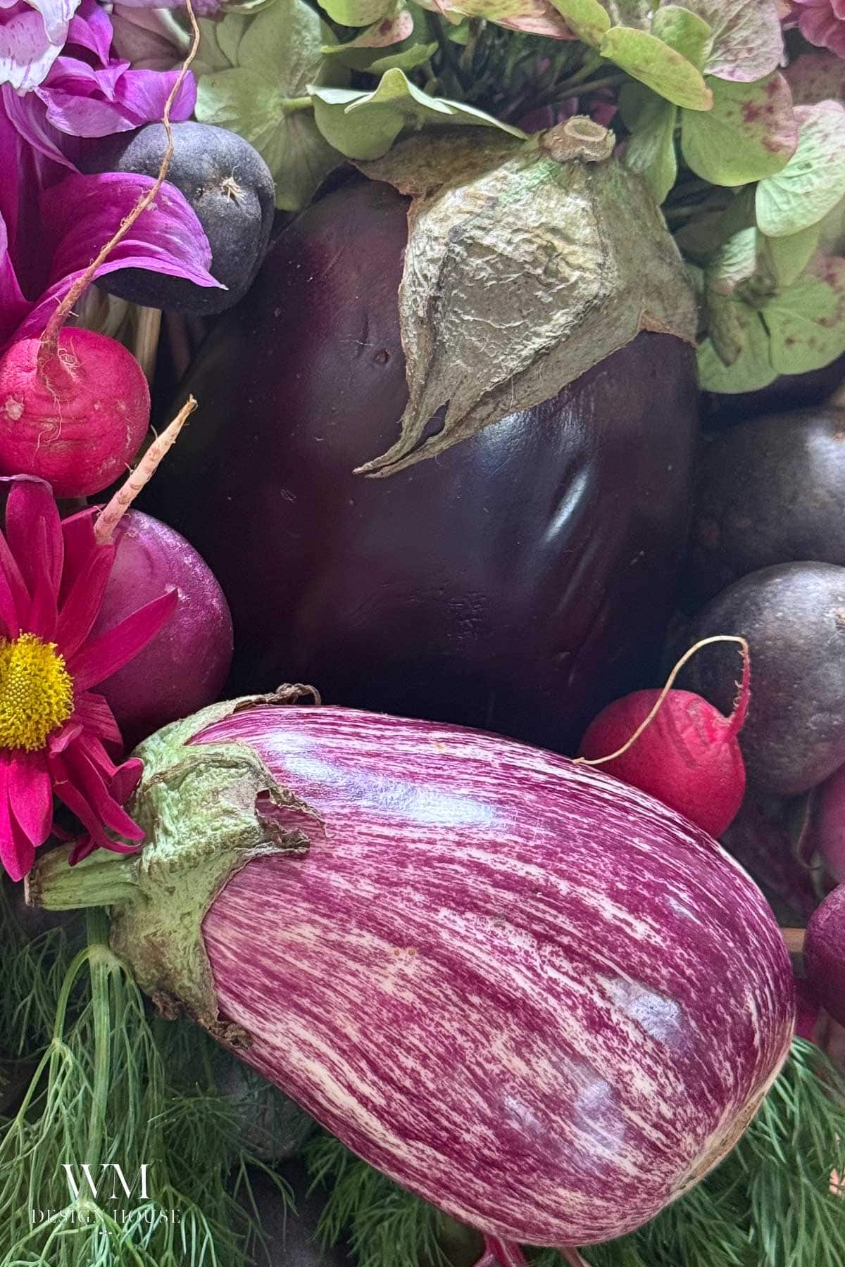 A close-up of a vibrant vegetable-centerpiece displays assorted vegetables, including a purple-striped eggplant, a dark purple eggplant, radishes, black potatoes, and flowers beautifully arranged together.