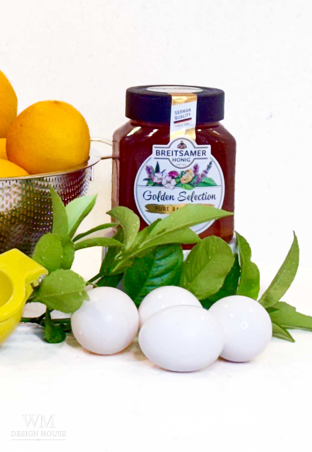 A jar of honey, three white eggs, a colander with lemons, and lemon leaves are arranged on a white surface—perfect ingredients for preparing homemade Meyer Lemon Curd or drizzling Salted Honey over your favorite desserts.