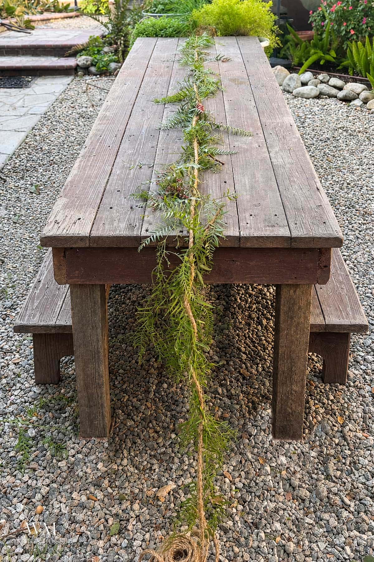 A rustic wooden table with matching benches sits on gravel, decorated with a simple greenery garland and twine along its center. Lush plants and stones surround the outdoor setting.